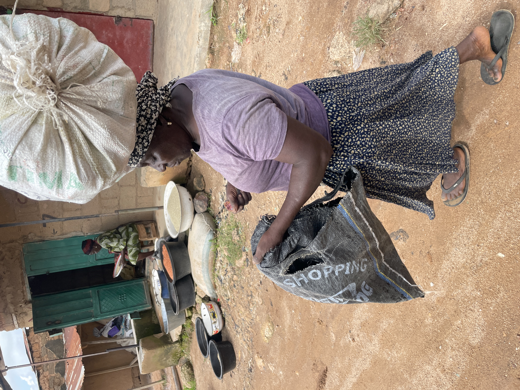 A woman in a purple t-shirt and a patterned blue skirt is walking on a dirt road in front of a building in Wang, Nigeria. She has a large sack of grain balanced on her head and is holding another sack in her hands. There are several buckets of grain and other goods in front of the building. The building has a red door and green shutters. There is a small child sitting inside the building. The area appears to be a market.
