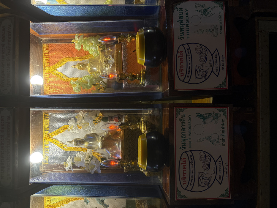 The image displays two adjacent, illuminated devotional display cases, likely situated within a temple or shrine in Ban Hin Sam Kon, Thailand. Both cases are constructed with dark wooden frames and glass fronts, each containing a seated Buddha statue.
The left display case features a translucent or light-colored Buddha statue, centrally positioned on an ornate golden pedestal. The interior background consists of a shimmering silver fabric panel, topped with a golden-yellow fringed valance. Two bouquets of white, artificial flowers are placed symmetrically on either side of the statue. In front of the Buddha, two electric 'flame-effect' lamps, emitting an orange-red light, stand on golden bases. Below this case, on a dark wooden shelf, a black offering bowl with a yellow slotted lid is visible. A rectangular white sign with a red border is affixed to the shelf, bearing Thai script and the English text "NIGHT WEDNESDAY," along with graphic representations of currency and symbolic Buddhist imagery.
The right display case holds a golden Buddha statue, similarly seated on an elaborate golden pedestal. Its interior background is an orange-gold patterned fabric panel, also crowned with a golden-yellow fringed valance. Two bouquets of light green/yellow artificial flowers flank this statue. Identical electric 'flame-effect' lamps, glowing orange-red, are positioned in front. Below this case, a black offering bowl with a yellow slotted lid is present. An accompanying white sign with a red border displays Thai script and the English text "THURSDAY," alongside currency graphics and symbolic Buddhist figures, including a standing Buddha. The environment beyond the cases appears dimly lit.