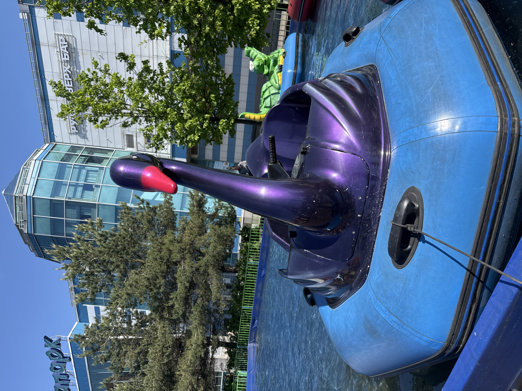 A purple swan-shaped boat is parked in a pool of water on a sunny day. The boat has a blue inflatable surround. In the background, there are buildings, trees, a green crocodile, and a fence. The building on the left has a sign that says "KIDBURG" in English. The other building has text in Russian "ВЕРХ БАР." The location is likely an amusement park in an urban environment in Unknown, Russia.