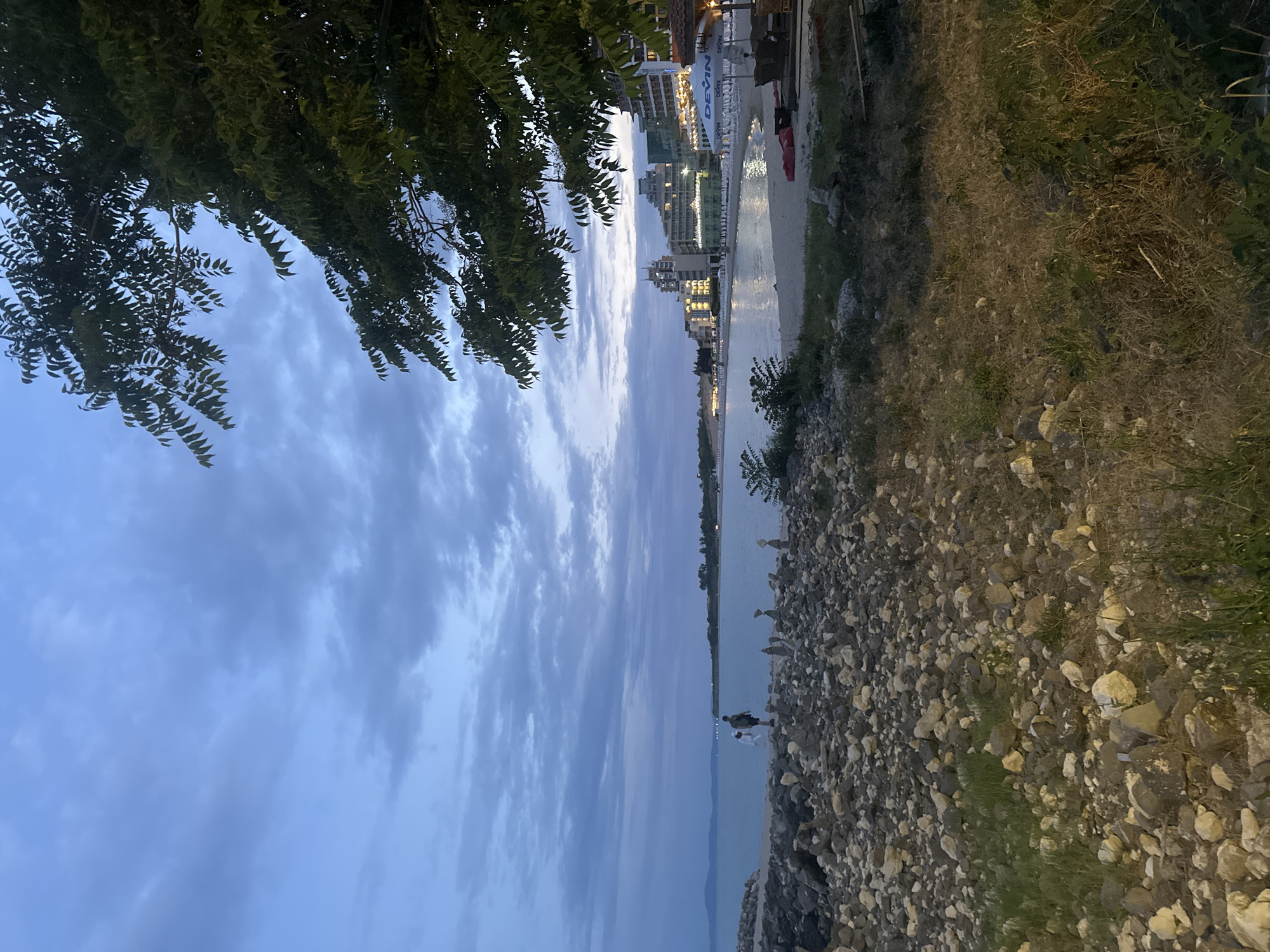 The image depicts a scenic view of a coastline with buildings in the distance. The scene is characterized by a body of water, a rocky shore, and a patch of dry grass and vegetation. On the left side of the image, a leafy tree partially obscures the sky, which is filled with soft, diffused clouds, suggesting it is either dawn or dusk.
In the water, there appear to be two people, one standing and one crouching, near the shore. The buildings in the background are illuminated, indicating that it is likely evening. The architecture suggests a resort or urban area along the coast. The overall atmosphere is serene, with the soft lighting and the gentle lapping of water contributing to a tranquil mood. The rocky shore is densely packed with stones of varying sizes. There is a sandy area visible between the rocks and the water. The dry grass and vegetation on the right side of the image suggest a dry climate or season.
On the buildings in the distance, there is visible text on what appears to be large white banners or signs. One sign clearly reads "DEVIN". The overall impression is a peaceful evening on a Bulgarian coastline, possibly in Nesebar, with a mix of natural beauty and human development.