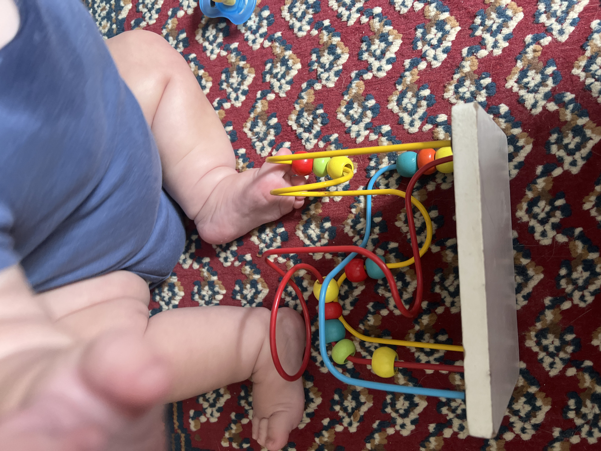 A close-up, high-angle shot shows a baby's legs and feet on a red patterned rug. The baby is wearing blue shorts. A wooden bead maze toy with red, yellow, blue, and green beads is positioned in the foreground, partially beneath the baby's feet. The toy has a white wooden base and colorful wire loops holding the beads. The rug has a repeating pattern of geometric shapes in dark red, blue, and white. To the far right, a blue pacifier is visible on the rug. The background is blurred, but appears to be a soft, light-colored surface, possibly a floor or a blanket.