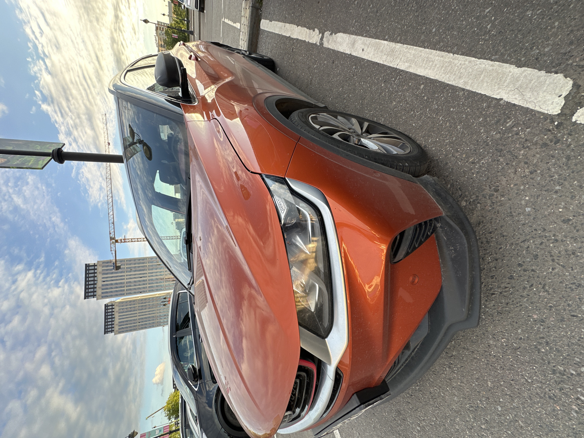 An orange-copper colored crossover SUV is parked on an asphalt surface under a partly cloudy sky. The photograph is taken from a low angle, focusing on the front-right side of the vehicle. The car's distinctive headlight, front grille with red accents, and part of the bumper are clearly visible. A multi-spoke wheel is seen on the right side of the car, positioned on the asphalt.
In the background, tall, modern buildings with numerous windows rise against the horizon. A large yellow construction crane is visible, towering over some of the buildings, indicating ongoing development in the area. The sky shows scattered, wispy clouds, suggesting a bright day. White parking lines are painted on the asphalt road surface, where other darker-colored vehicles are partially visible parked behind the main subject car. A black pole with an illegible sign stands to the left of the car. The overall lighting suggests it is daytime, likely late morning or afternoon. No identifiable text is clearly visible on any object. The scene depicts an urban environment, consistent with a city like Moscow, Russia.