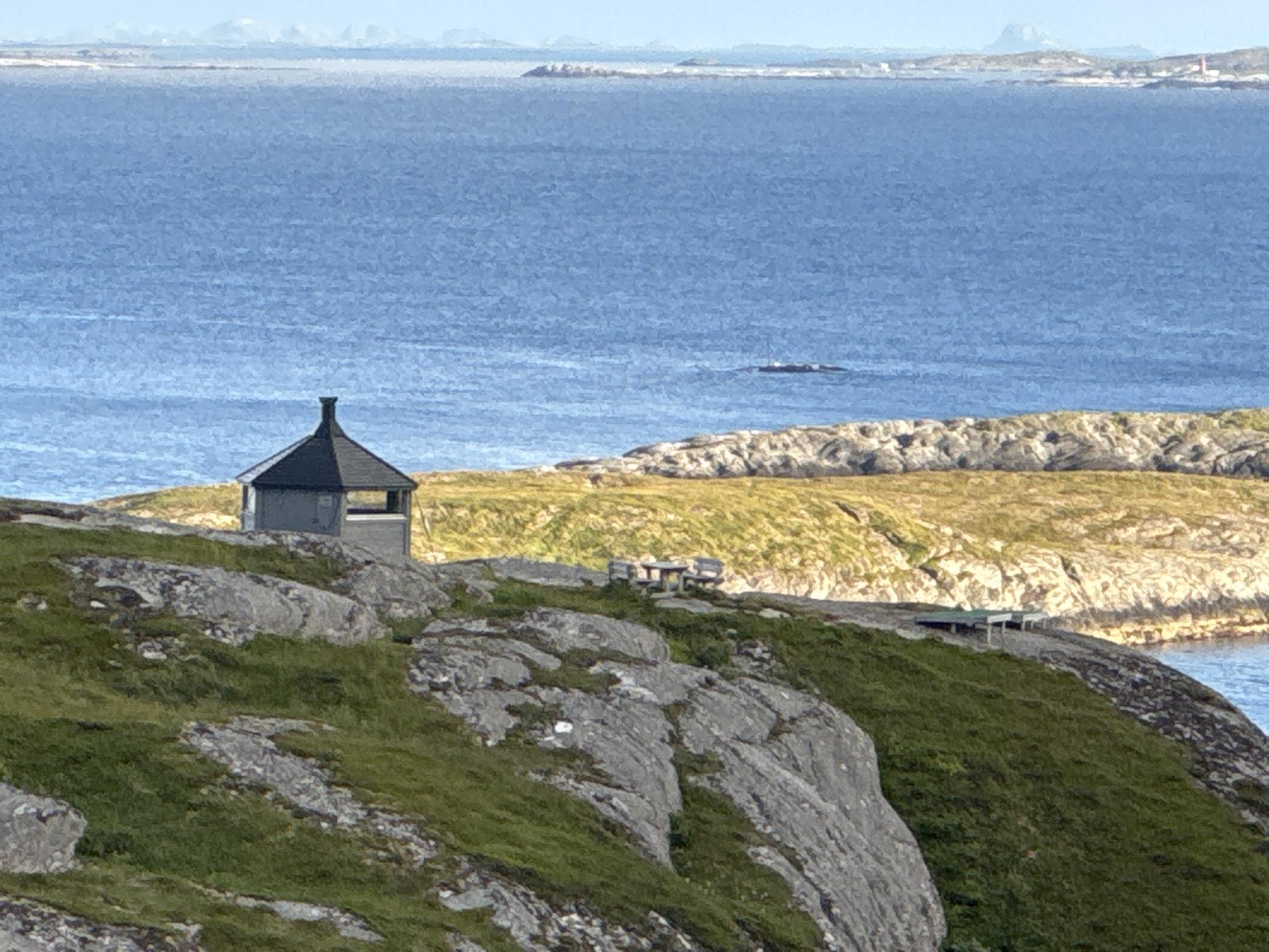 A small, dark gray, octagonal structure with a pointed roof sits on a rocky, grassy outcrop overlooking a body of blue water. In the foreground, the land is covered with green grass and large gray rocks. In the middle ground, a picnic table with two benches is visible on a grassy area. Further in the background, more rocky islands and distant mountains are visible across the water under a bright sky. The scene is of a coastal environment on Sleneset, Norway.