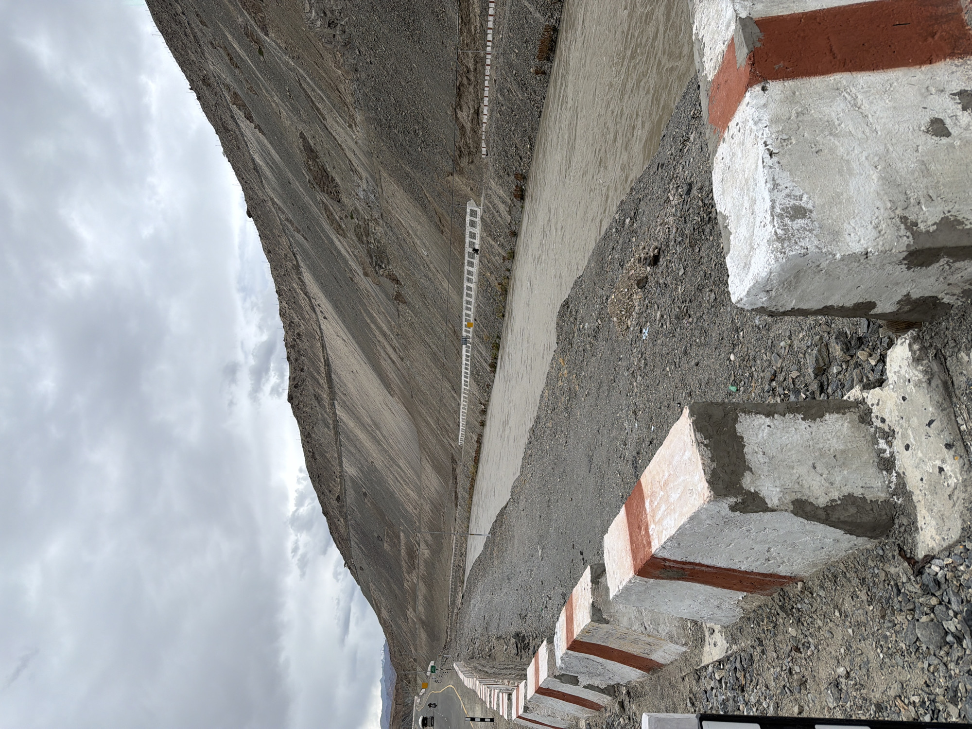 This is a medium shot, eye-level, outdoor photograph taken in Chumkar, India. The scene depicts a wide, muddy river flowing through a valley flanked by steep, barren, mountainous terrain under a cloudy sky. In the foreground, a road barrier made of concrete blocks painted with red and white stripes lines the edge of the road. The road itself is paved and curves to the left, with a few vehicles, including motorcycles and what appear to be trucks, visible in the distance. The mountainous slopes are covered in loose gravel and rock, with some evidence of man-made structures, such as retaining walls and possibly a few buildings or encampments, built into the slopes. The overall impression is one of a remote and rugged landscape.