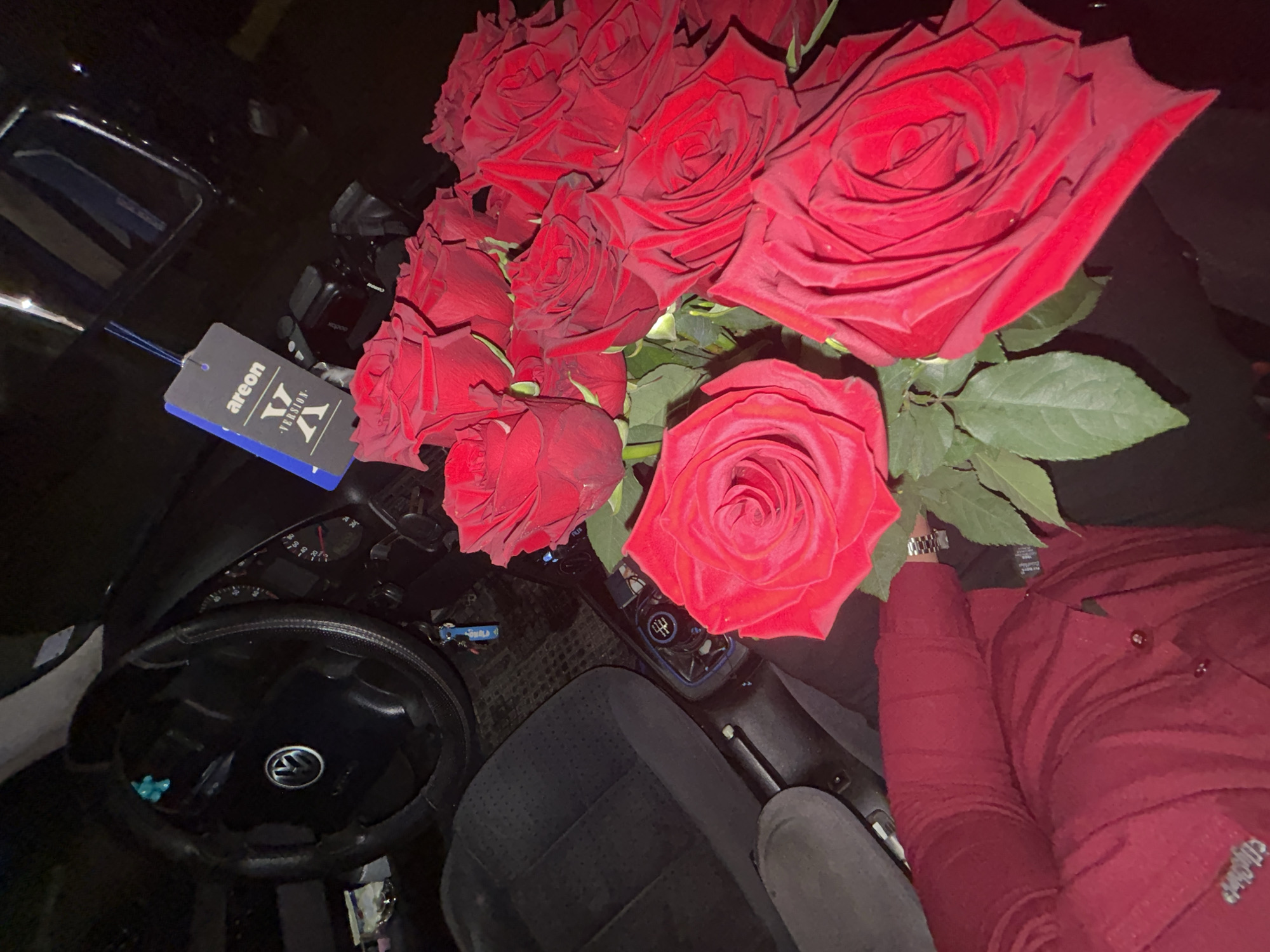 The image shows the interior of a Volkswagen car, specifically the driver's seat and dashboard. A large bouquet of red roses sits in the passenger seat. An "Areon" air freshener hangs from the rearview mirror. A person's arm and part of a burgundy-colored shirt are visible on the right side of the frame. The vehicle's steering wheel is prominently displayed. There is no information about location in the city of Zlatograd, Bulgaria.