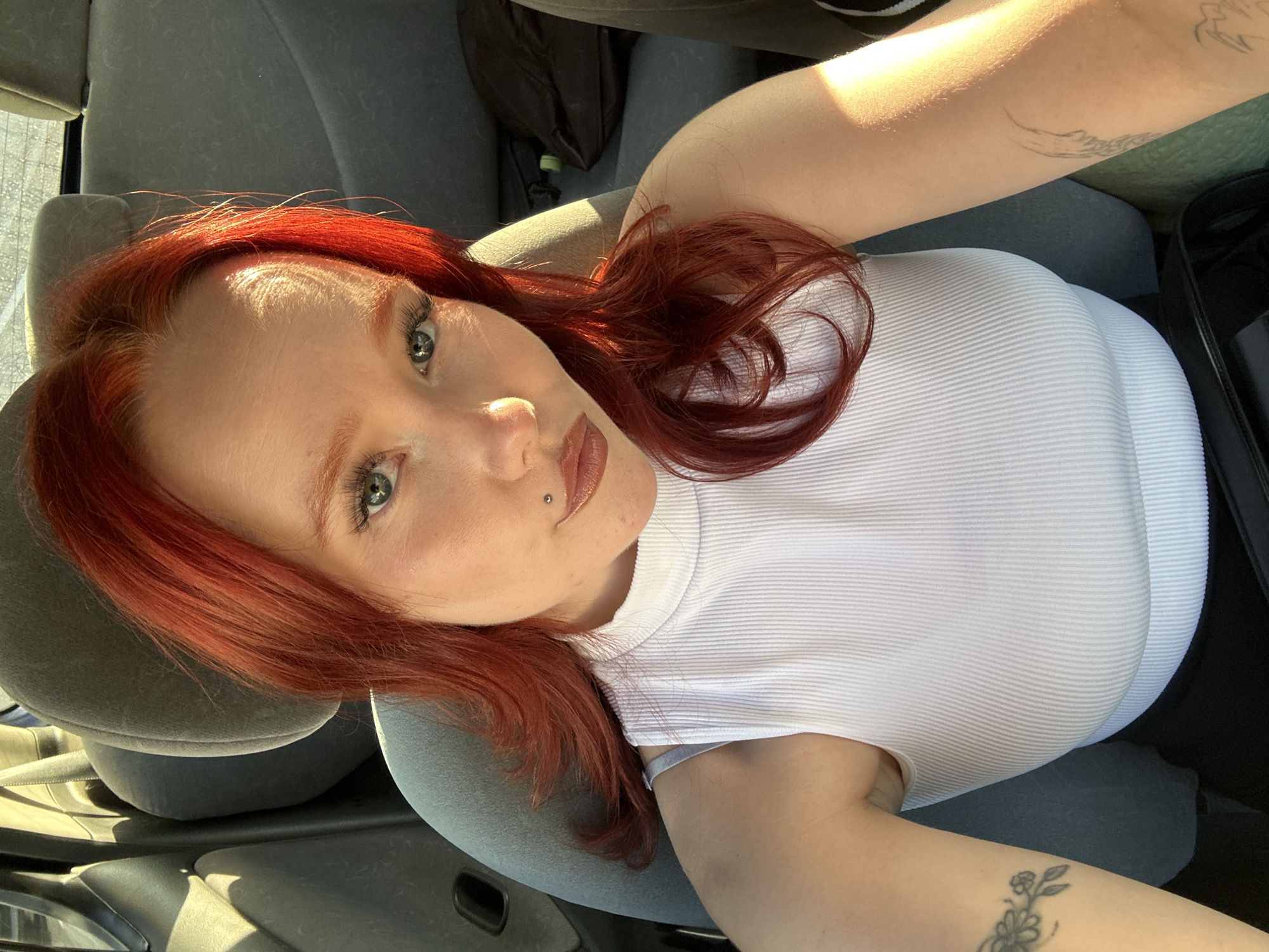 A young woman with vibrant red hair, wearing a white ribbed tank top, is captured in a close-up, selfie-style photograph. Her eyes, a striking blue-green, gaze directly at the camera. A small silver lip ring is visible on her lower lip. Tattoos adorn her arms, one appearing to be a line drawing of waves on her upper arm, and another a floral design on her forearm.
The woman is seated in what appears to be the driver's seat of a car, with the grey car seat visible around her. Sunlight streams in from the left side, casting a warm glow and highlighting the texture of her hair and skin. The background suggests an indoor setting, possibly a vehicle interior, with a dark object possibly a bag, and part of the dashboard visible. The lighting indicates it is daytime, with strong sunlight creating bright highlights and some shadows. The overall mood appears relaxed and candid. No text is visible in the image.
