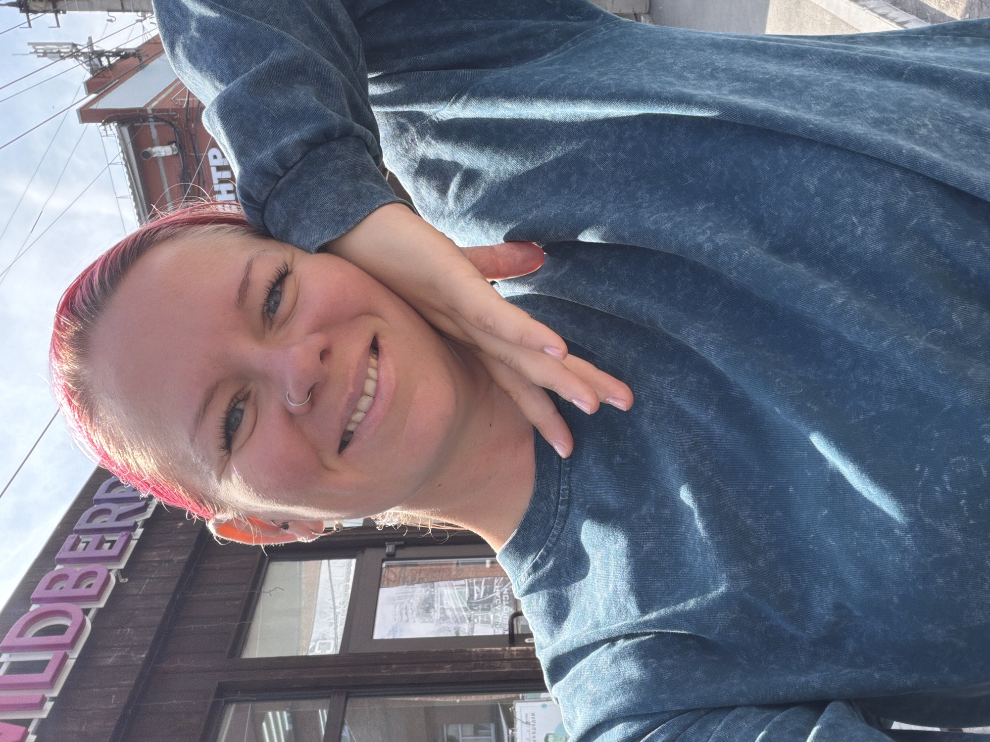 A woman with pink-dyed hair is taking a selfie outdoors. She is wearing a teal, tie-dyed sweatshirt and has a nose ring and several earrings. Her left arm is bent, with her hand resting on her cheek, and her right arm is behind her head, obscured by her hair. She is smiling at the camera. Behind her, a building with the sign "Wildberries" is visible. Another building with Cyrillic lettering is further in the background, along with a clear sky. The scene appears to be in a commercial area, likely in Votkinsk, Russia, given the Cyrillic text.