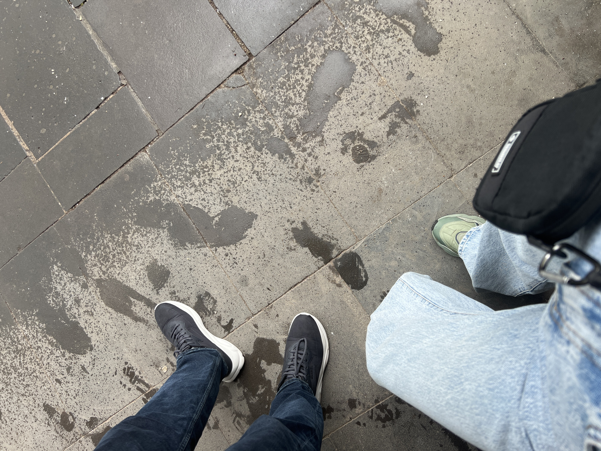 The photograph depicts a street scene in Tbilisi, Georgia. The ground is paved with dark grey stone tiles, some of which show signs of wear and cracking. There are water stains or footprints scattered across the pavement. Two pairs of legs and feet are visible. One person wears dark blue jeans and dark sneakers with white soles. The other person wears light blue, baggy jeans and one green sneaker is visible. A black bag with a silver buckle is visible hanging on their person. The perspective is from an elevated viewpoint, likely taken by one of the individuals depicted.