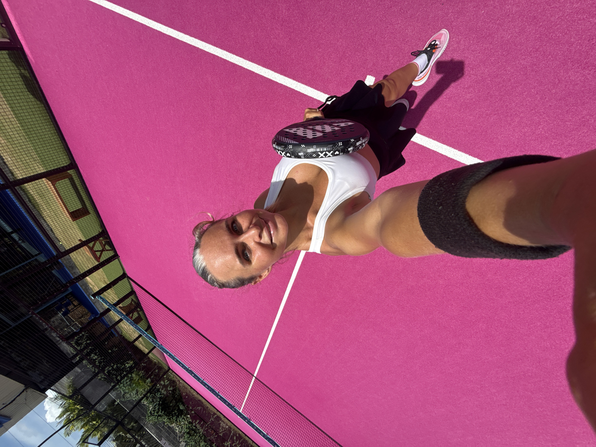 A woman with blonde hair pulled back and grey streaks, wearing a white sports bra and a black skirt, stands on a bright pink padel court. She is holding a black and white padel racket in her right hand. The court has white lines. The background shows a fence, greenery, and a blue sky with some clouds. The woman is smiling at the camera, taking a selfie. This appears to be an outdoor padel court in a sunny location.