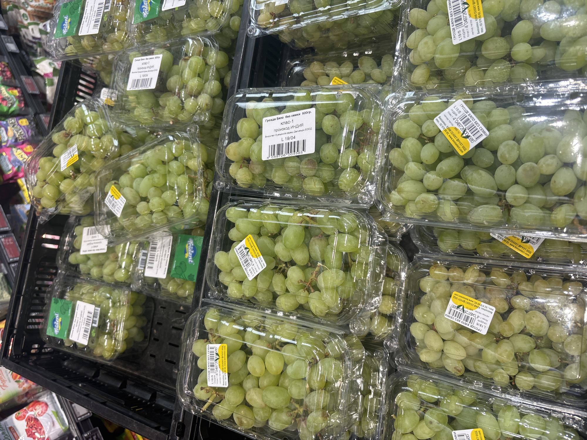 The image shows a stack of clear plastic containers filled with green grapes, arranged within a black crate in a supermarket. Each container has a label with product information in Bulgarian and English, including the name "Грозде Бяло без семки" (White Seedless Grapes), weight, and country of origin (India). The supermarket is located in Plovdiv, Bulgaria, as indicated by the brand "КОНИ СЕЛЕКТ ФРУХТ ООД" on the labels, a Bulgarian fruit company. Several containers also display the "Trident" brand. The grapes are the primary focus of the image, presented as merchandise for sale.