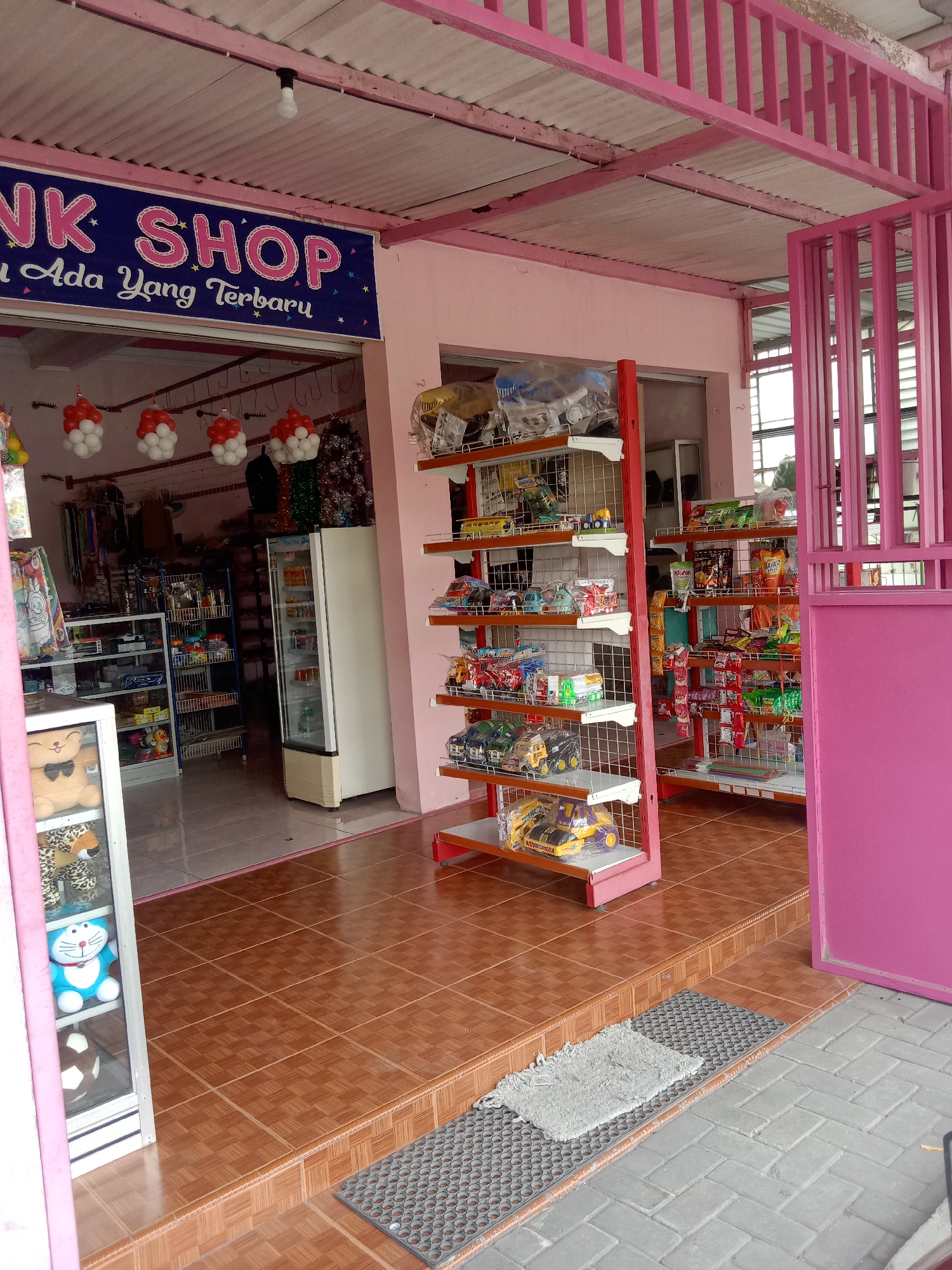 The image depicts the exterior of a small shop with a pink facade and a pink metal gate. A blue banner above the entrance reads "NK SHOP" in large pink letters, with smaller text below that appears to be in Indonesian. Red and white balloon decorations are hung from the ceiling inside the shop. Shelves displaying various toys and snacks are visible both inside and outside the shop. A white refrigerator is positioned inside the shop, and a display case with plush toys, including a blue Doraemon plush, is located near the entrance. The ground in front of the shop is tiled with brown squares, and a gray mat is placed on the tiled steps leading into the shop. The overall impression is of a small, brightly colored convenience store or toy shop. The shop appears to be located in a residential or commercial area in Mayangkawis, Indonesia, as suggested by the architecture and store type.