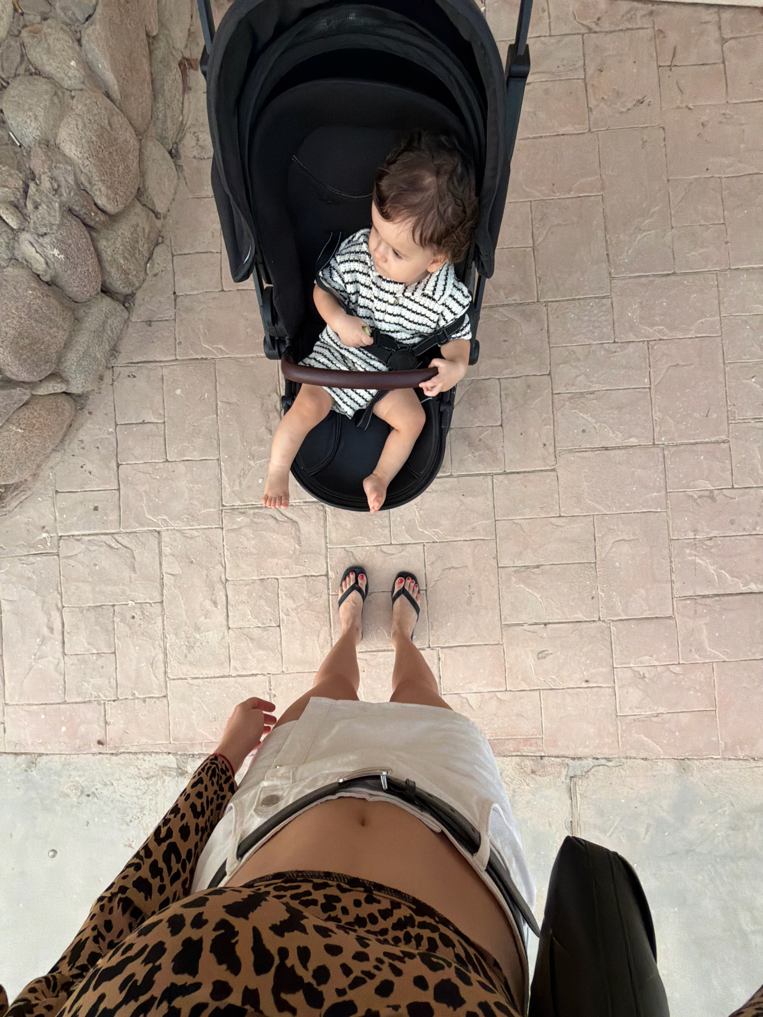 An overhead, medium shot captures a woman and a baby in a stroller on a paved outdoor patio. The woman, positioned at the bottom of the frame, wears a leopard-print long-sleeved top and a white skirt. Her legs are visible from the knees down, and she is wearing black flip-flops. To the left of her, a large black stroller is occupied by a baby dressed in a black and white striped outfit. The baby's legs are visible as they rest on the stroller's footrest. The ground is covered in light brown rectangular pavers. To the left of the stroller, a wall made of rough, grey stones is visible. The scene is likely located in Sharm El Sheikh, Egypt, given the architectural style and warm-toned paving stones commonly found in resort areas.