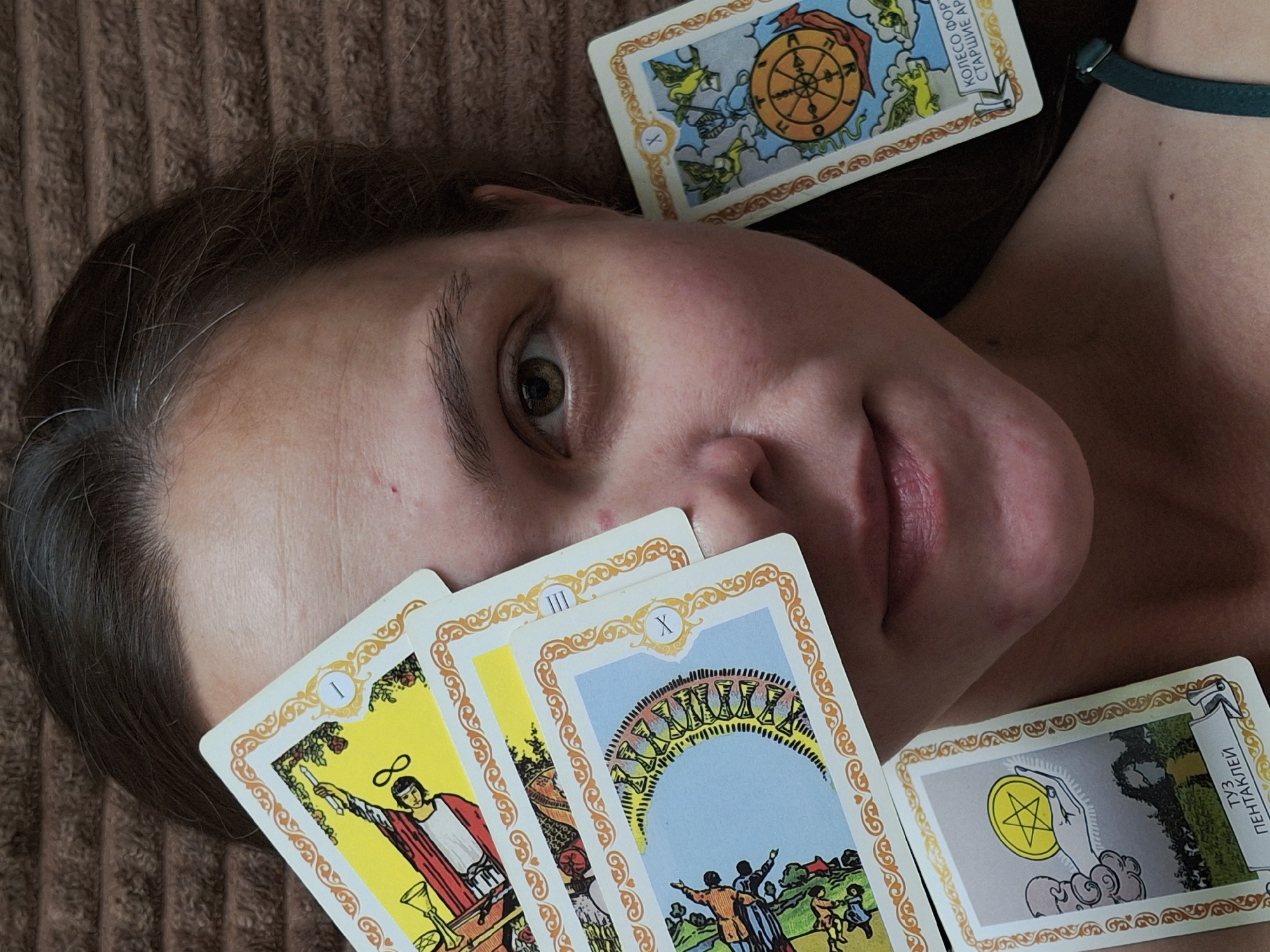 The image features a close-up, overhead view of a person lying on their back on a brown, textured surface, possibly a blanket or rug. The person's face is angled towards the viewer, with their right eye and part of their mouth visible. They have dark hair with some grey strands. Scattered across the person and the surface are several tarot cards.
One card, the Wheel of Fortune, is positioned above the person's forehead. To the left of the face, fanned out, are at least three other tarot cards. The visible cards include the Ace of Wands, the Three of Cups, and the Ten of Cups, all from the Major Arcana. Another card, the Ace of Pentacles, is visible on the right side of the image, near the person's shoulder. The cards depict various scenes and symbols typical of tarot decks, with intricate illustrations and borders. The person is wearing a green strap, possibly part of a garment, around their upper arm.
The lighting appears to be soft and diffused, suggesting an indoor setting, possibly during the daytime. The overall mood conveyed is one of introspection or perhaps a consultation of fate, given the presence of the tarot cards. There is no discernible weather or strong location cues beyond the indoor setting.
Visible text on the cards includes Roman numerals (I, III, X), the phrase "КОЛЕСО ФОРТУНЫ" (Wheel of Fortune) and "СТАРШИЕ АРКАНЫ" (Major Arcana) on the Wheel of Fortune card, and "ТУЗ ПЕНТАКЛЕЙ" (Ace of Pentacles) on the Ace of Pentacles card. The image was likely taken in Chaikovsky, Russia, based on the provided context.