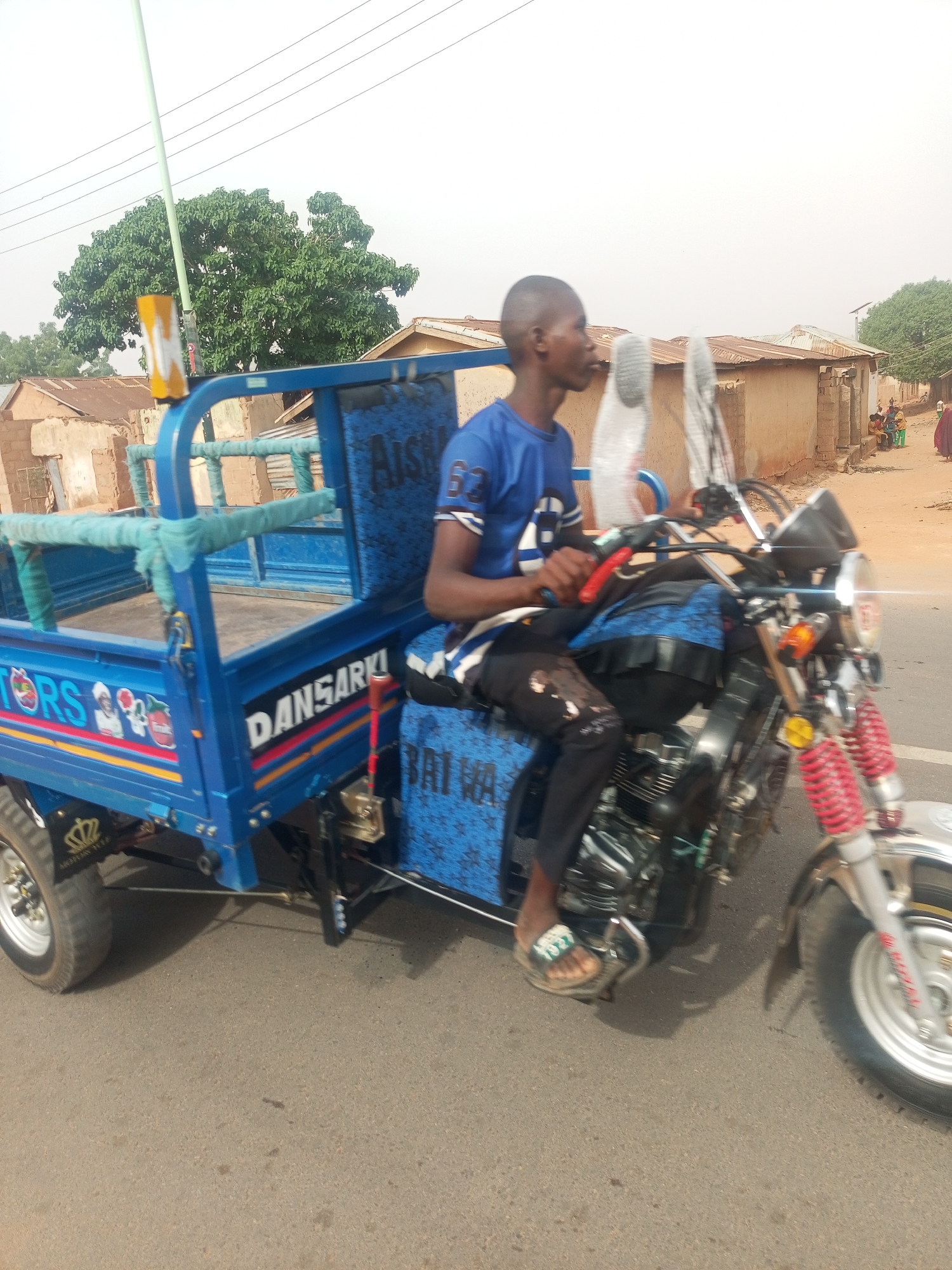 A young man in a blue t-shirt and dark pants sits on a blue motorized tricycle with a cargo bed. The tricycle has chrome accents and colorful decals. The location appears to be Tempure, Nigeria, as indicated by the backdrop of simple, earth-toned buildings and a few people in the distance. The tricycle is on a paved road. Overhead power lines are visible. The sky is overcast.