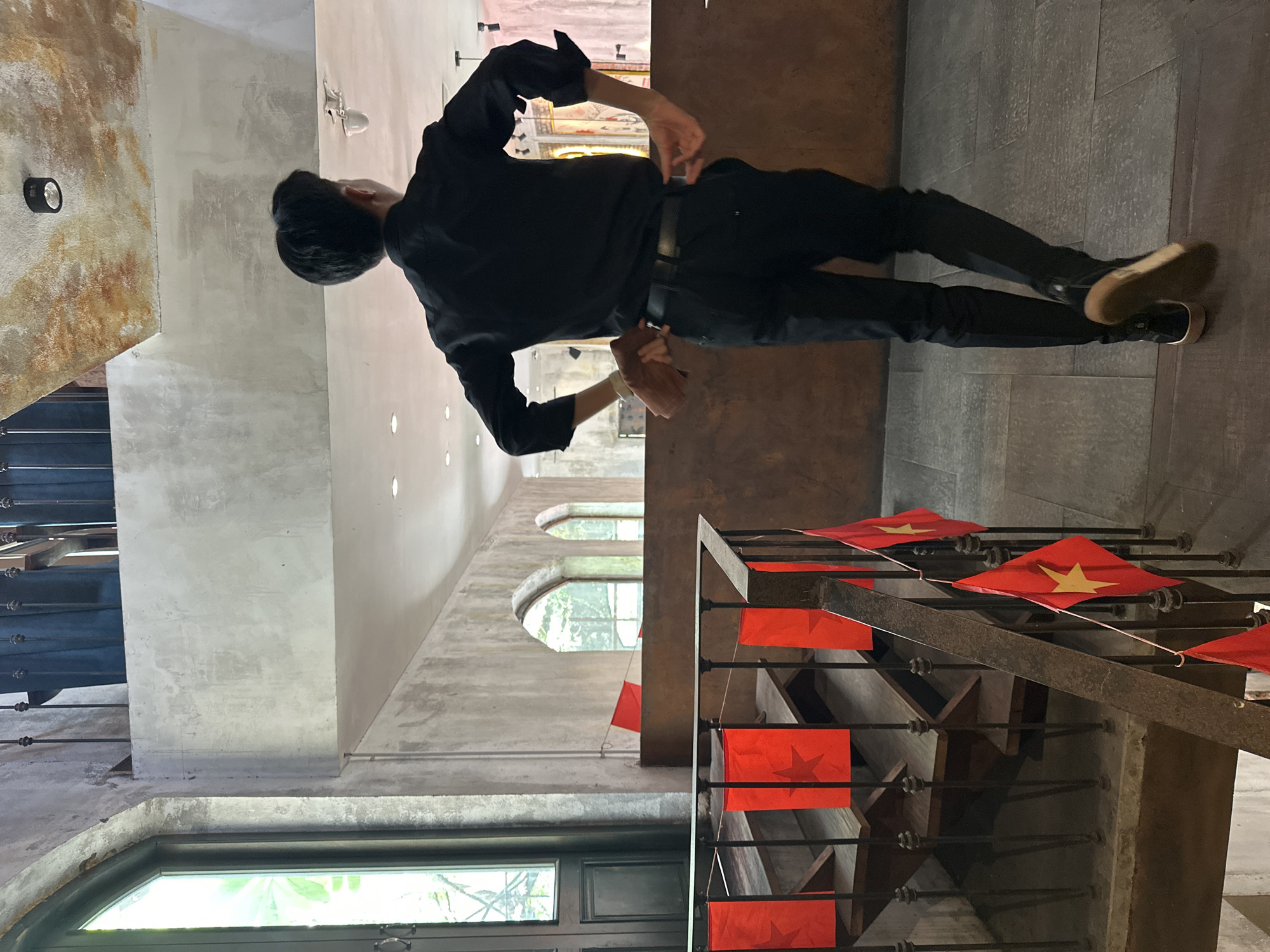 The image depicts an indoor scene within an establishment, likely a multi-level cafe or restaurant in Vietnam, given the presence of Vietnamese flags. The specific location within the city of Đà Nẵng is not discernable from the visual information provided.
In the foreground, a person, viewed from behind, is walking away from the camera towards the center-right of the frame. They are dressed in a long-sleeved black shirt, black trousers, and dark sneakers with light brown soles. Their right hand holds a folded brown object, possibly a menu or cloth, while their left hand is near their hip, appearing to adjust their clothing or access a pocket. Their dark hair is neatly cut short.
To the left, a wooden staircase with dark metal railings descends from an upper level. Small red flags adorned with a yellow star (the national flag of Vietnam) are strung along the metal railing of the staircase. Additional red flags, some appearing as simple rectangles, are attached to the wall further up the stairs. The walls throughout the space are textured and distressed, resembling concrete or stucco in shades of grey and beige.
On the left side of the image, a tall, dark-framed window allows natural light to enter, revealing greenery outside. In the background, on the upper level, several arched windows with dark frames also showcase lush outdoor foliage. A low, dark brown partition or counter separates the foreground area from the space with the arched windows.
The ceiling is multi-tiered; a lower section directly above the person is smooth and light-colored with embedded spotlights, while an upper, higher section exhibits a mottled, textured surface with patches of rust-orange and grey, featuring a black track light fixture. The floor in the foreground consists of light grey rectangular tiles interspersed with darker wood-like strips. The overall aesthetic is rustic and industrial.