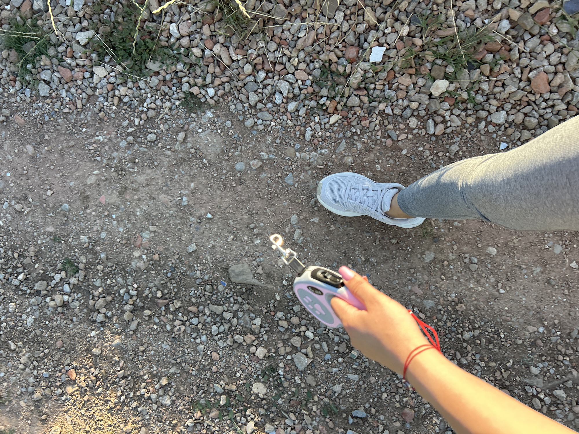 An overhead shot shows a person's legs and arm as they walk on a gravel path. The person is wearing gray athletic pants and white sneakers. Their right arm, adorned with a red string bracelet, is holding a retractable dog leash. The leash is pink and gray with a silver clip. The path is covered in small, varied-colored stones and dirt. Patches of dry grass and weeds are visible along the edge of the path. The lighting suggests it is daytime, possibly late afternoon, with the sun casting long shadows.