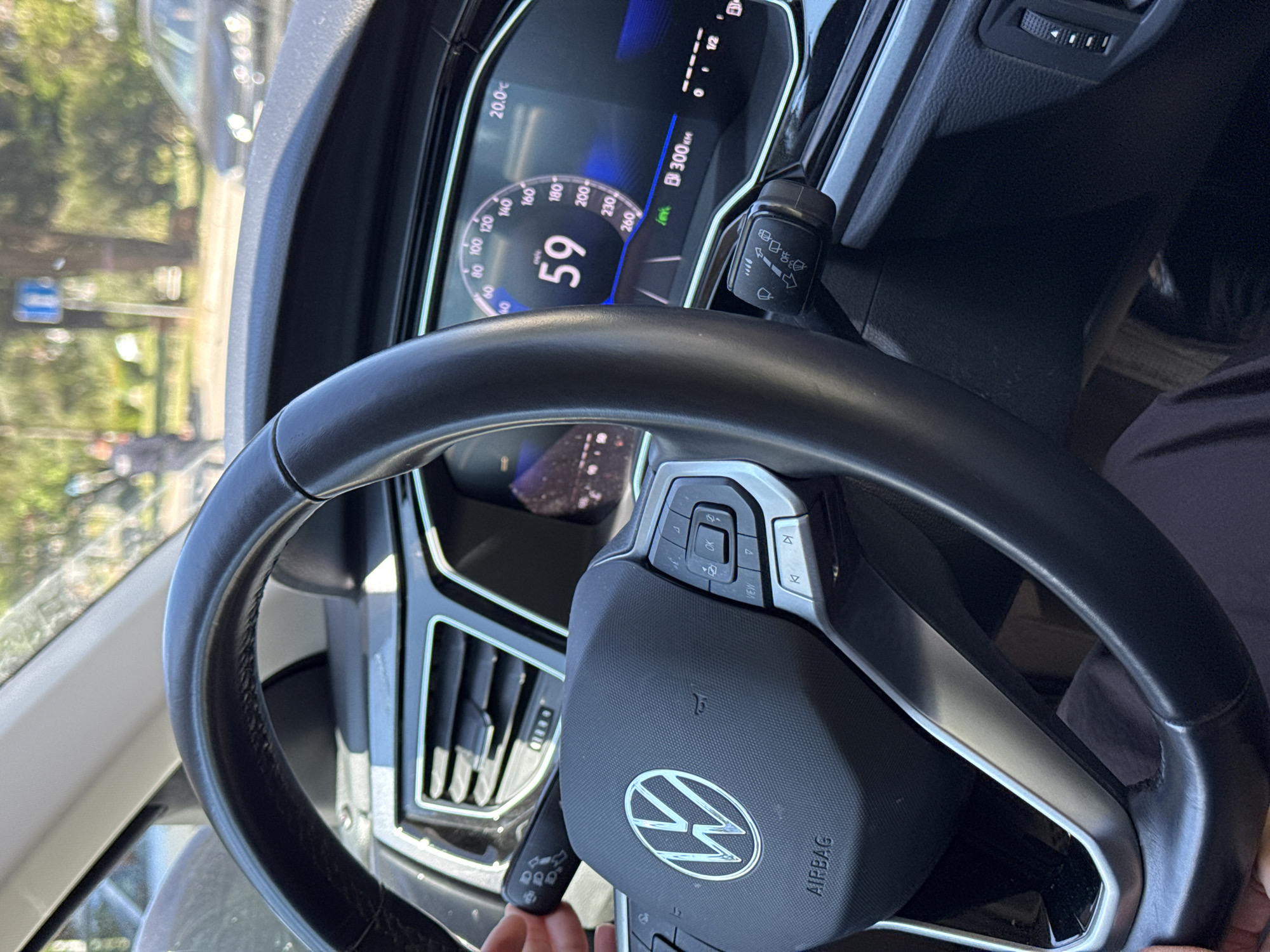 The image shows the interior of a car, focusing on the dashboard and steering wheel. The car is a Volkswagen, as indicated by the logo on the steering wheel. The speedometer reads 59, and the digital display shows 20.0°C. The car has traveled 300km. Outside the car, trees and a blurred building are visible. Given the limited visual context, it is impossible to definitively place the location in Budva, Montenegro. There is a hand visible at the bottom of the image, suggesting someone is driving the vehicle. Another person is in the passenger seat.