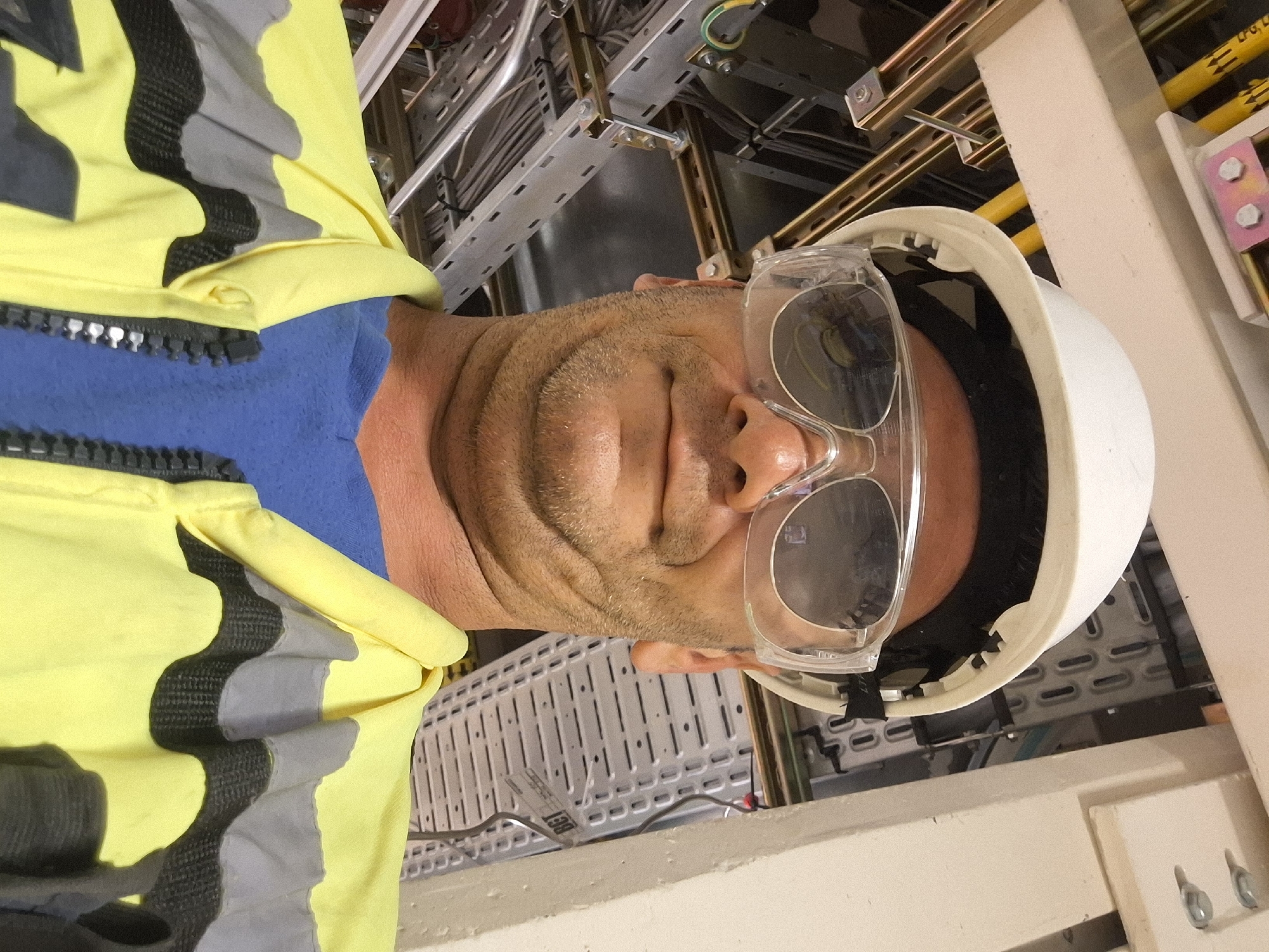 This image, taken in Kiryat Gat, Israel, shows a close-up selfie of an adult male wearing safety gear. The man has short, stubbly facial hair and is wearing a white hard hat and clear safety glasses. He is also wearing a bright yellow high-visibility vest over a blue t-shirt. The background reveals an industrial or construction environment with exposed electrical wiring, conduits, and metal framing structures. The lighting suggests an indoor setting, possibly a facility undergoing maintenance or construction.