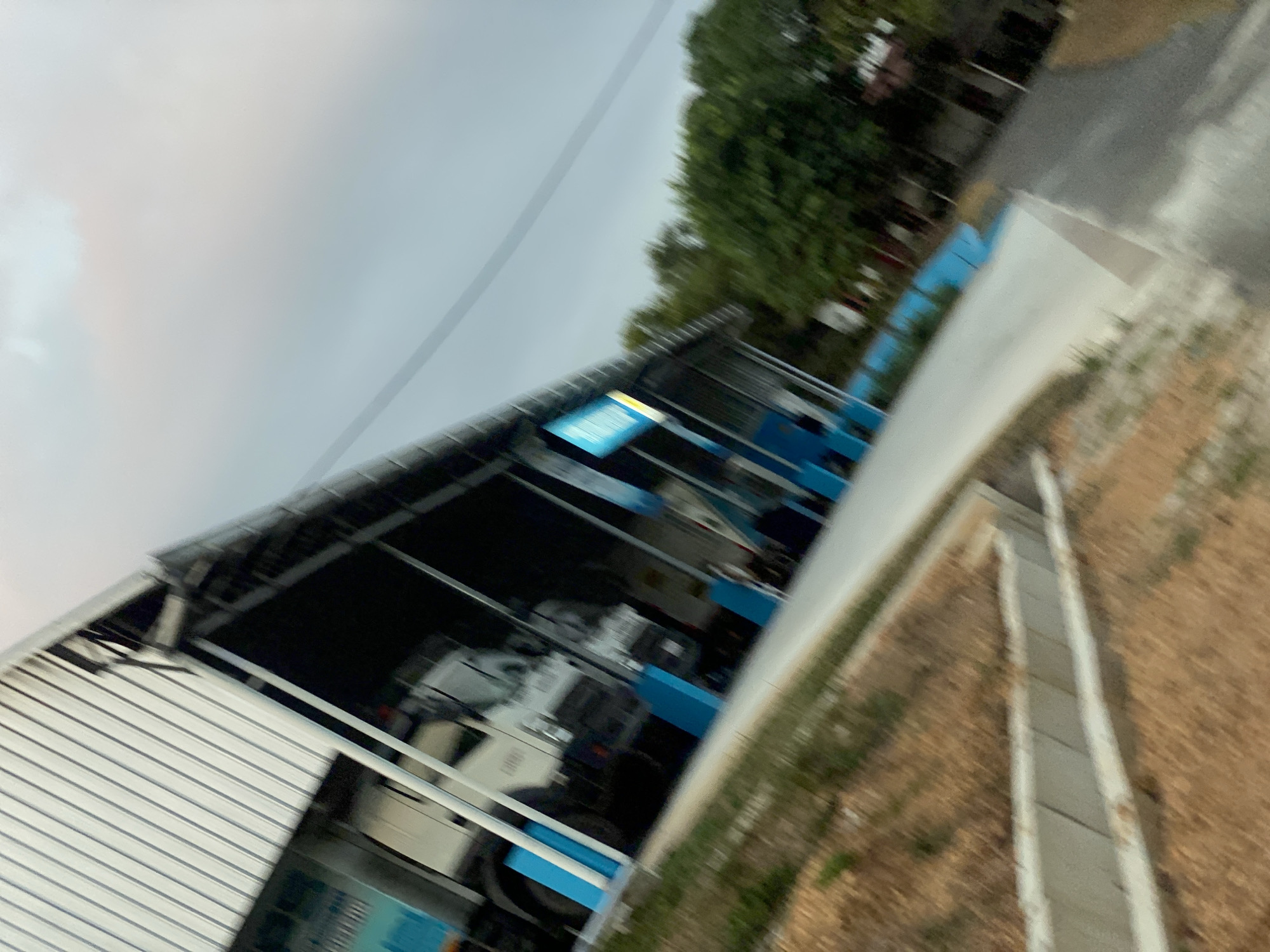 The image depicts an outdoor scene, likely in Banjarbaru, Indonesia. A white truck is parked under a covered structure with a corrugated metal roof. The structure appears to be a carport or a maintenance bay, with blue pillars supporting the roof. In the background, there are trees and a cloudy sky. The ground in front of the structure is a paved surface, and to the left, there is a sloped area with dry grass and a concrete embankment. The overall impression is of a functional, possibly industrial or commercial, setting. The image is captured at an angle, giving it a dynamic and slightly abstract quality.