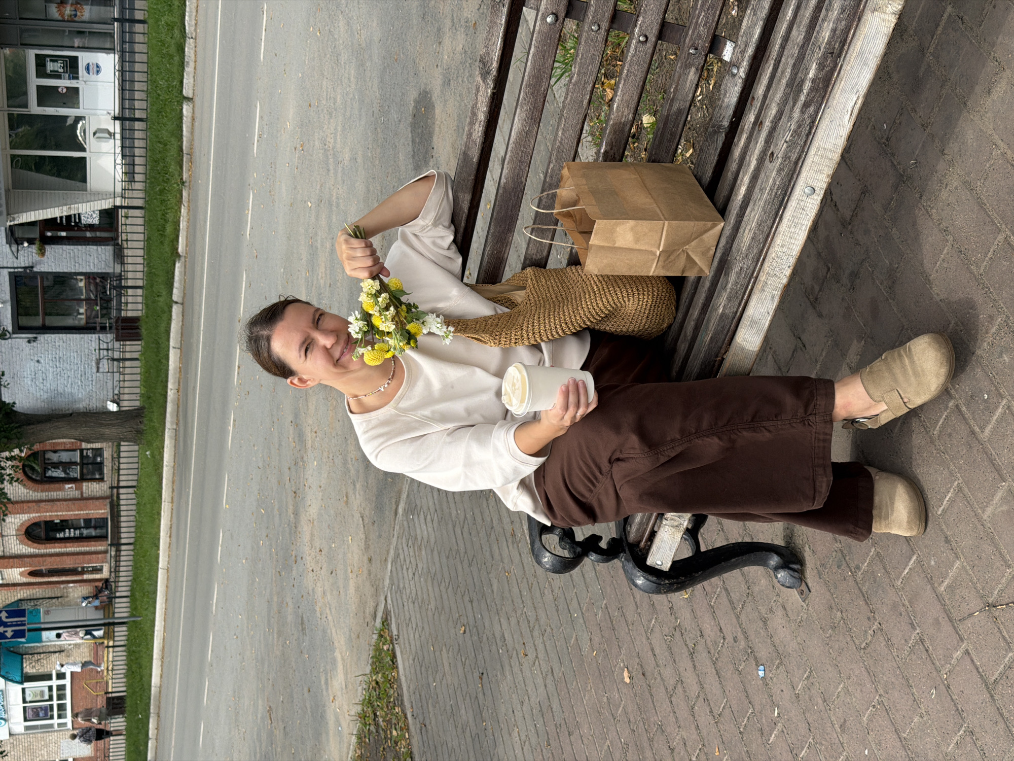 A woman sits on a park bench in Chaikovsky, Russia, holding a small bouquet of yellow and white flowers and a paper cup. She wears a cream-colored long-sleeved shirt and brown wide-leg pants, with tan slip-on shoes. Next to her on the bench are a woven straw bag and a brown paper shopping bag. The bench is made of wood and metal, situated on a paved walkway. In the background, a street is visible with cars and buildings. The environment appears to be an urban setting with trees and grass bordering the street. The woman is smiling and looking at the camera.