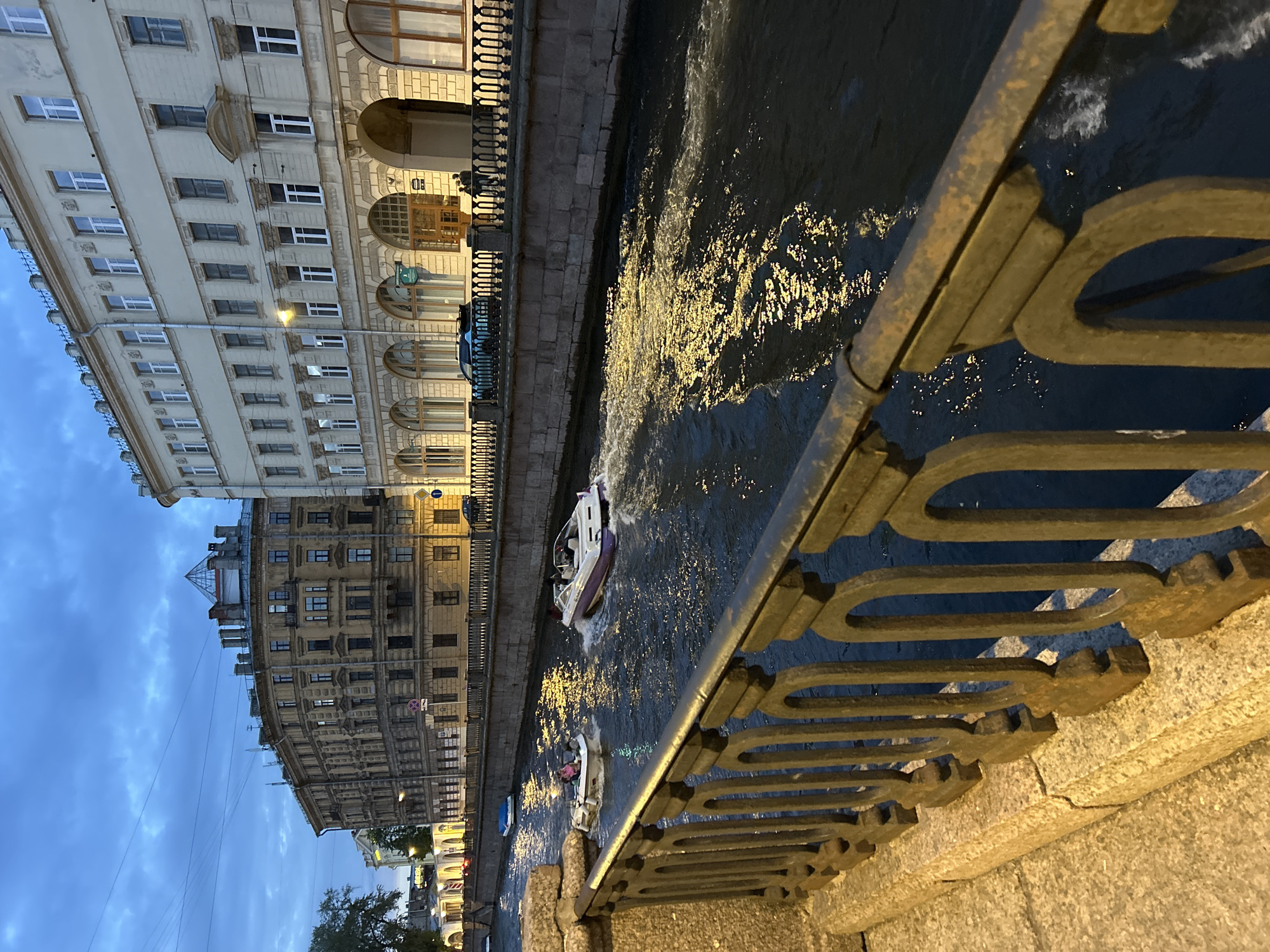 The image depicts a canal in a European city, likely at dusk or early evening. The canal is flanked by stone embankments and ornate metal railings in the foreground. Buildings with multiple stories and numerous windows line the canal on the right, their facades illuminated by streetlights. On the left, a tree and more buildings are visible, with some illuminated windows.
Several small motorboats are navigating the canal. One boat, with a purple and white hull, is moving away from the viewer, creating a wake. Another smaller white boat with two occupants is closer to the left bank, also in motion. The water in the canal reflects the lights from the buildings, creating a shimmering effect. The sky is overcast with a deep blue hue. The overall impression is of a picturesque urban waterway scene.