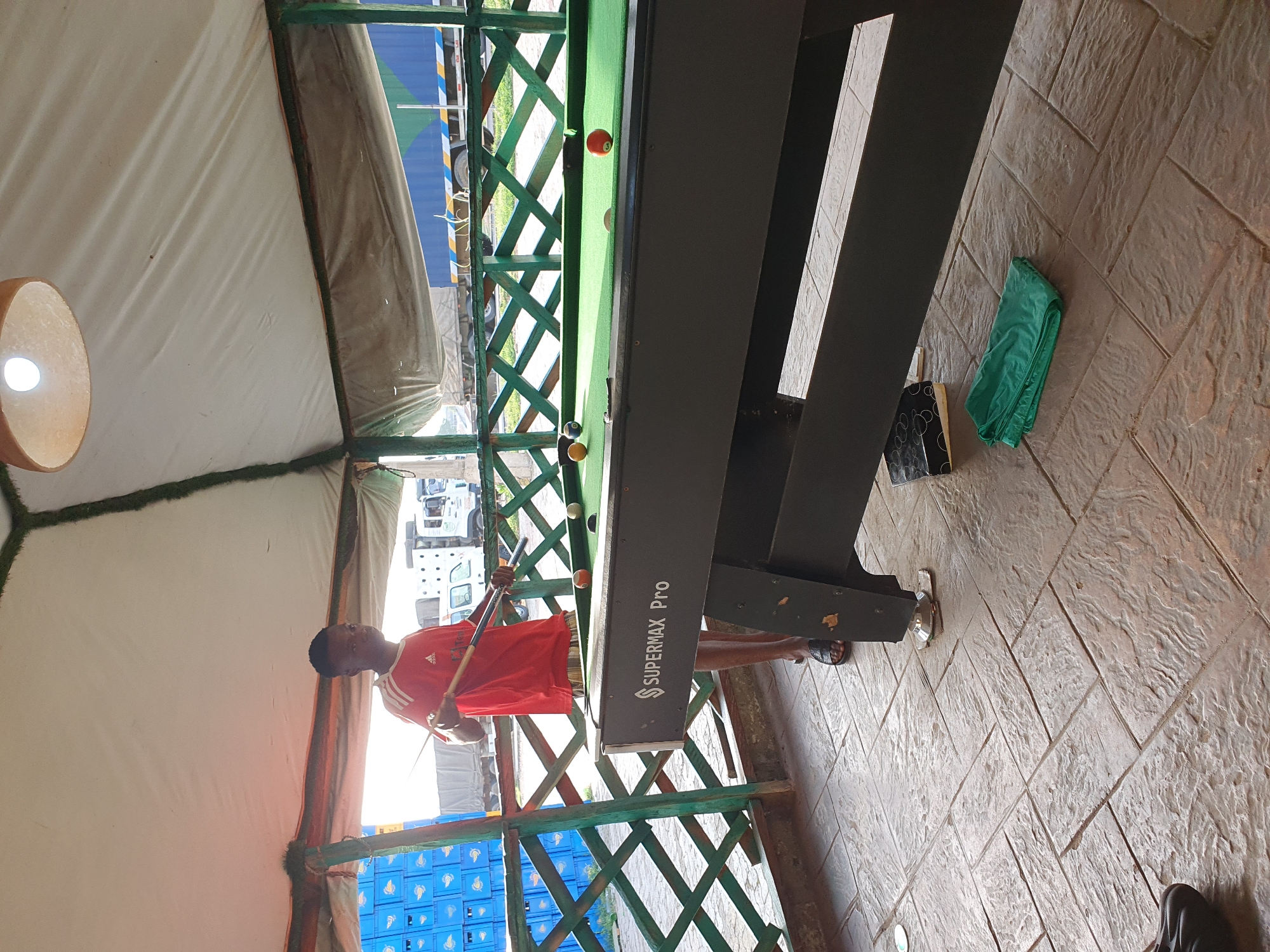 A person is playing pool in a covered outdoor area. The scene is outdoors, with a light-colored tent or awning overhead. The floor is paved with textured stones. A pool table, labeled "SUPERMAX Pro," is central to the image. Several pool balls, including an orange one with the number 3 and a yellow one with the number 1, are visible on the green felt of the pool table. The person, wearing a red shirt with white stripes on the sleeves and dark shorts, is holding a pool cue and appears to be taking a shot. The person is facing away from the camera, towards the pool table. In the background, there are stacks of blue crates and some vehicles, suggesting an area with commercial activity. The lighting suggests it is daytime. The overall atmosphere seems casual.