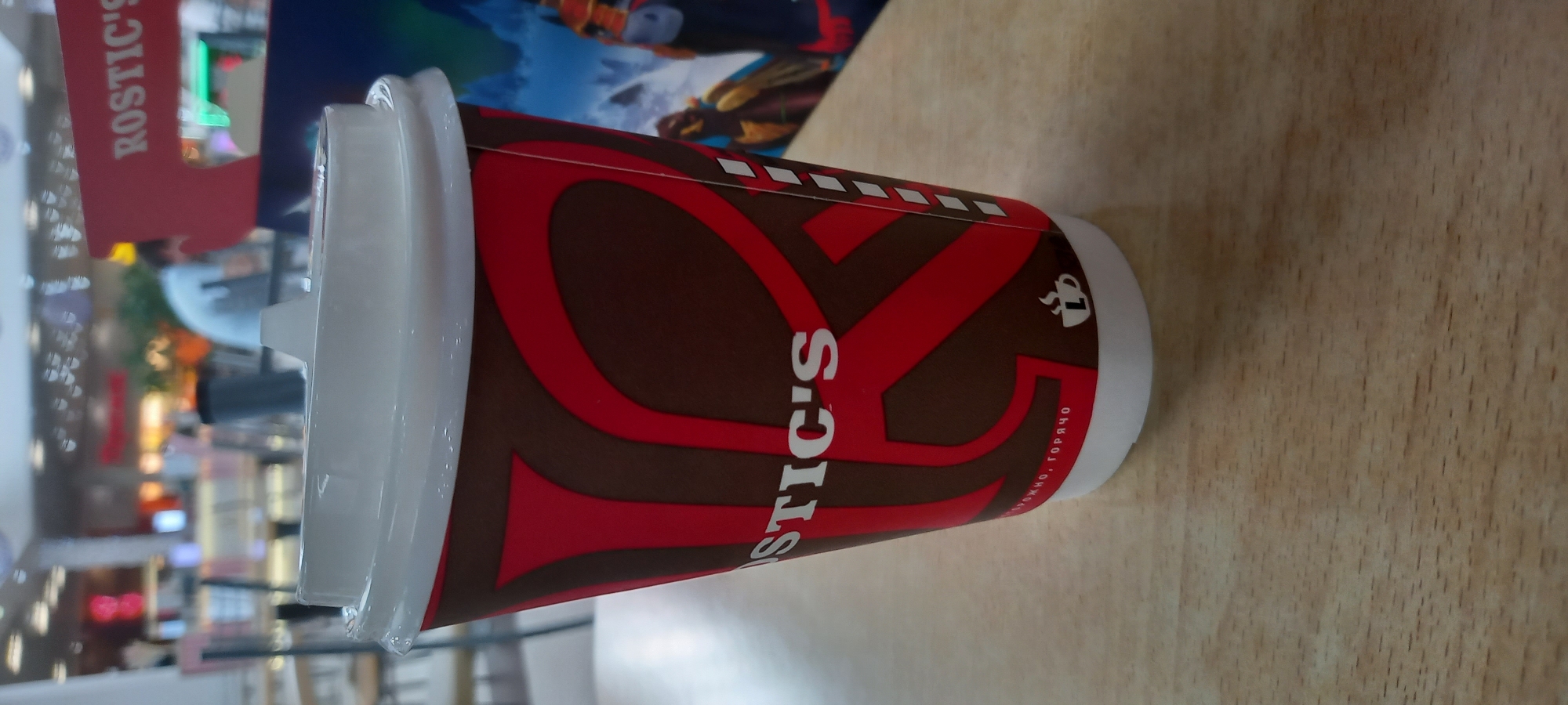The image depicts a partially visible Rostic's coffee cup placed on a light-colored surface with a wood grain texture. The cup is white at the top and bottom, with a brown and red Rostic's logo printed on the side. The background shows a blurry interior scene with other objects and figures, suggesting a public space. Based on the logo and general ambiance, this image was likely taken in a Rostic's establishment located in Krasnodar, Russia.