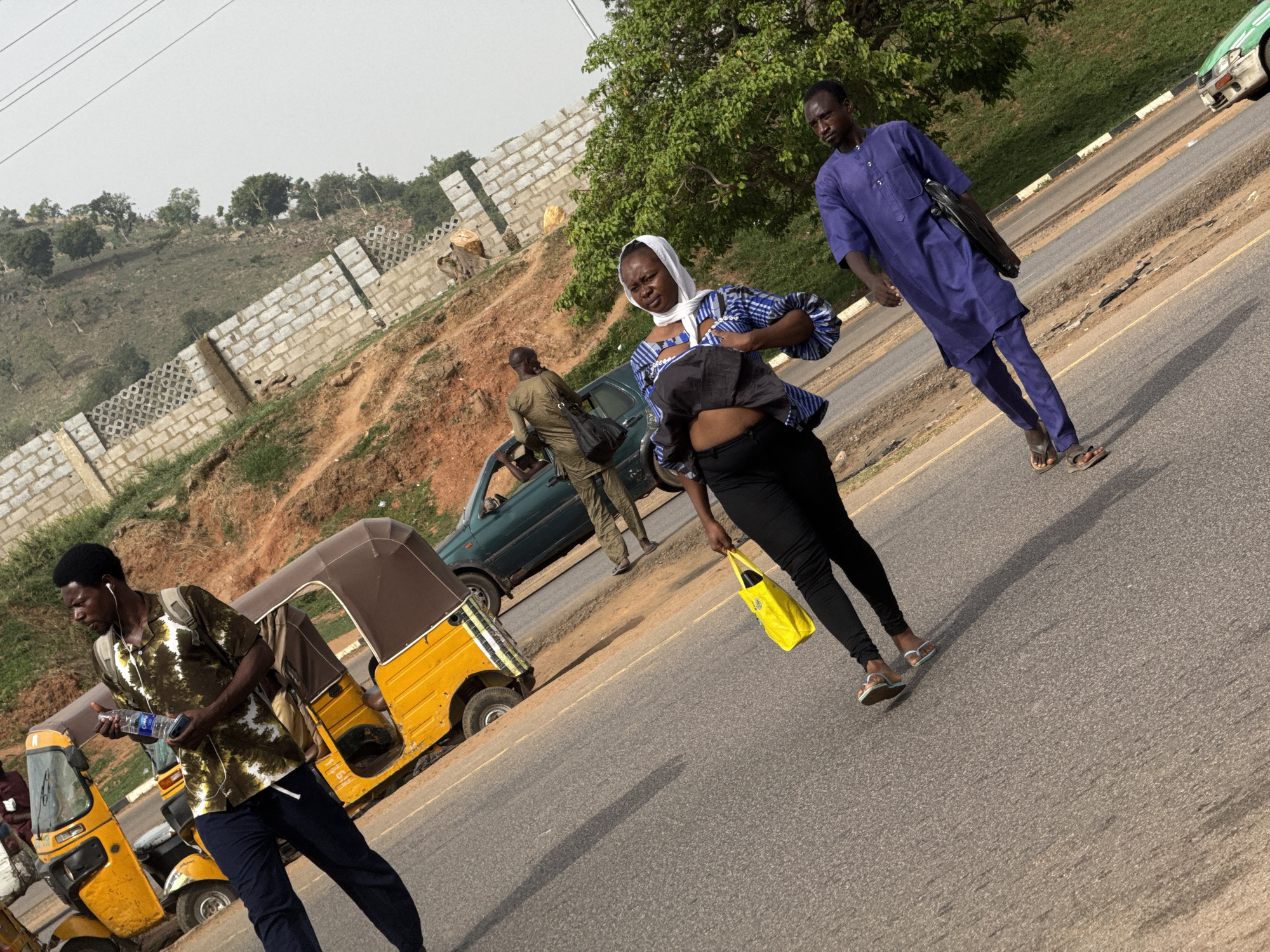The image depicts a street scene in Abuja, Nigeria. A main road runs diagonally through the frame, with several people walking along its edge. In the foreground on the left, a man in a patterned shirt and dark pants is holding a water bottle and appears to be listening to headphones. Behind him are several yellow "Keke Napep" tricycles.
Further along the road, a woman wearing a headscarf and a patterned blouse is walking. She is carrying a yellow bag. Next to her, a man in a purple outfit and sandals is walking.
In the background, there is a parked green car and another man in a green outfit is standing near the car. Behind them, there is a hillside with a partial concrete block wall and trees.