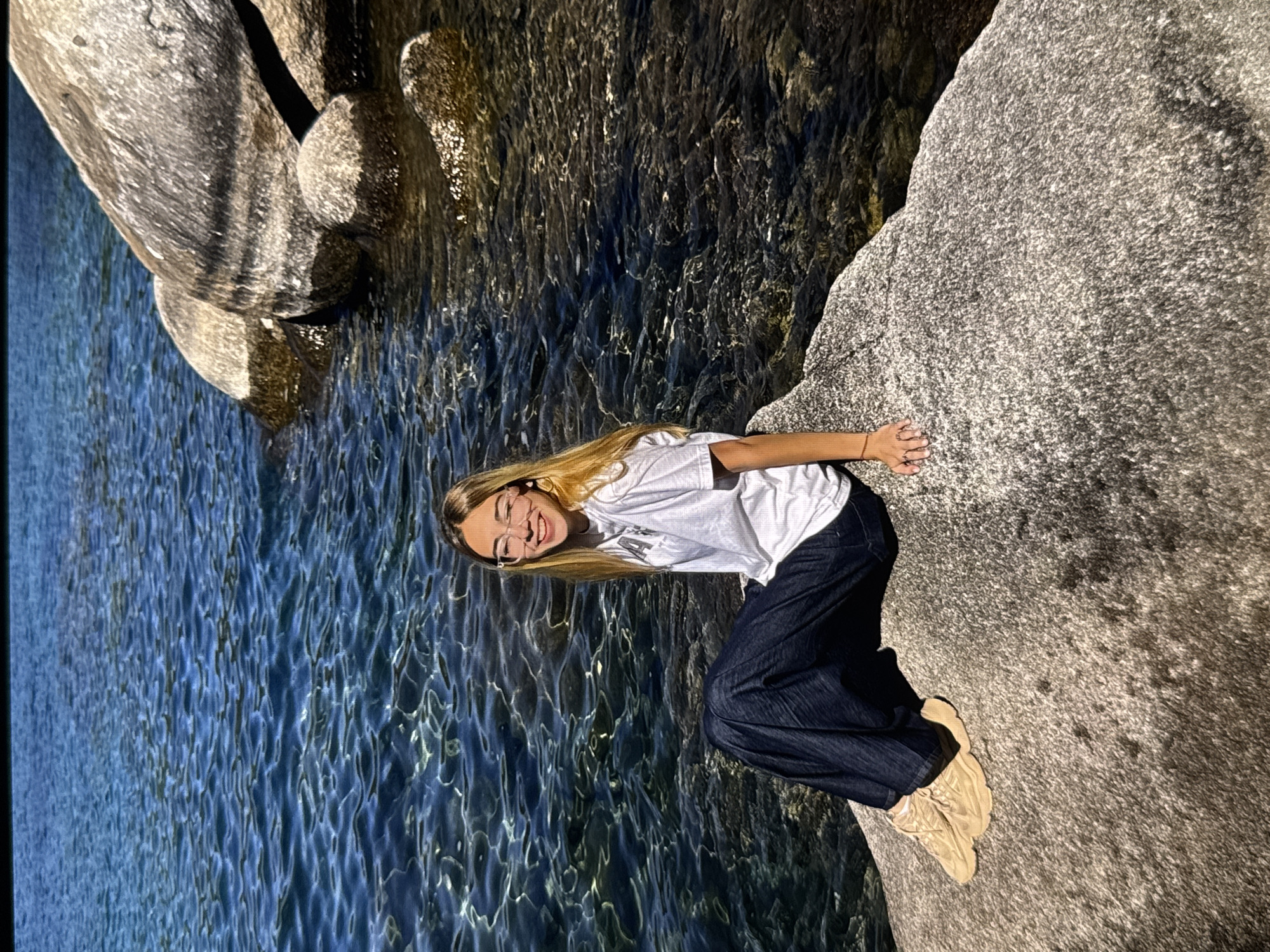 A young woman with blonde hair and glasses is sitting on a large rock next to a body of water. She is wearing a white t-shirt and dark pants. The water is clear and blue, with sunlight reflecting off the surface. There are also some large rocks in the water and along the shore. The image appears to have been taken outdoors on a sunny day. The location is not specified in the image.