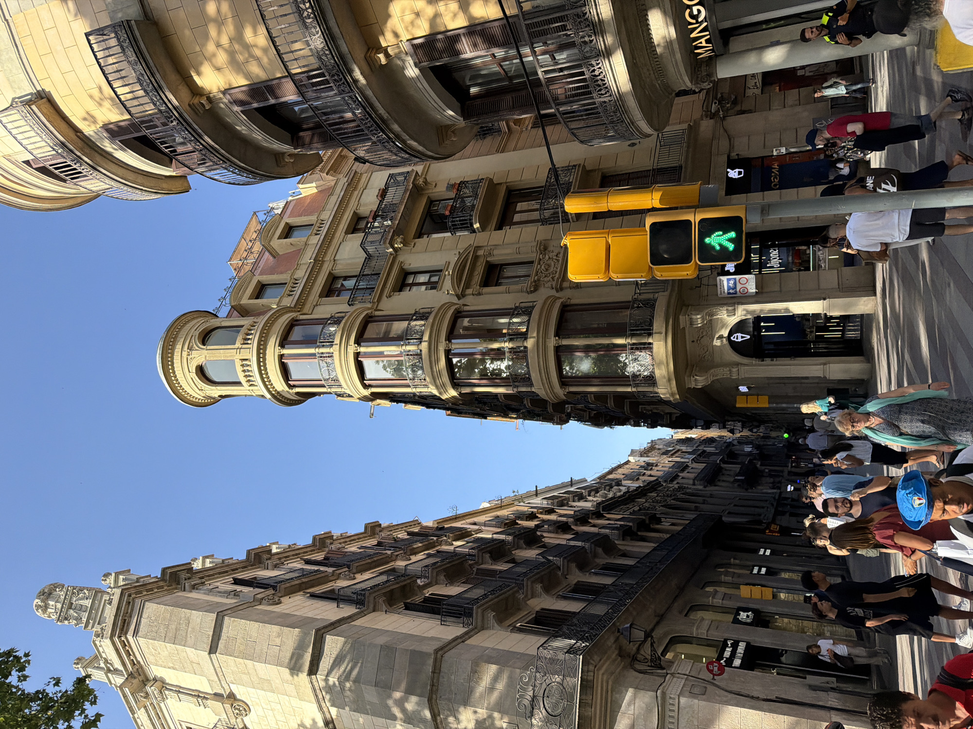 A wide-angle, eye-level outdoor shot captures a bustling street scene in Barcelona, Spain, on a clear, sunny day. The architecture is a mix of ornate, older buildings with stone facades, decorative balconies, and large windows, juxtaposed with modern shop fronts. The street is paved with stone, and the buildings are tightly packed, creating a canyon-like effect.
In the foreground, several people are walking on the sidewalk. A man in a red shirt and a blue beret is prominent, facing left. To his right, a woman in a patterned dress and a light blue scarf walks towards the viewer. A man in a white t-shirt and shorts is visible, facing left, and a woman in a dark dress is to his left.
A bright yellow traffic light stands prominently on the right side of the image, showing a green pedestrian signal, indicating it is safe for people to cross. One of the shops visible has a prominent "MANGO" sign.
The background shows more of the narrow street lined with buildings, with a clear blue sky overhead. The sunlight casts distinct shadows on the buildings and the street, highlighting the architectural details and the activity of the pedestrians.