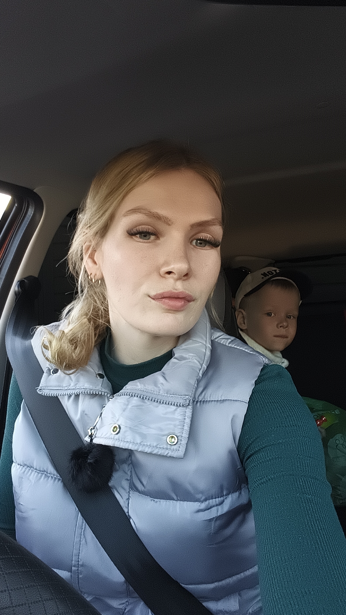 Inside a car, likely in Русский Вожой, Russia, a woman takes a selfie, with a young child visible in the background. The woman, positioned in the foreground and wearing a seatbelt, has blonde hair tied back, prominent eyelashes, and a neutral expression with a slight smirk. She wears a light grey or blue puffy vest over a dark teal ribbed long-sleeved top. A black fluffy pom-pom charm is attached to the zipper of her vest.
Behind her, in the back seat, a young child looks towards the camera with a calm or serious expression. The child wears a light-colored baseball cap with partial dark text, possibly "MOT," and a white collar or neck warmer. A green object, possibly a car seat or bag, is visible behind the child. Natural light illuminates the scene, suggesting it is daytime. The car's dark headliner is visible at the top of the image.