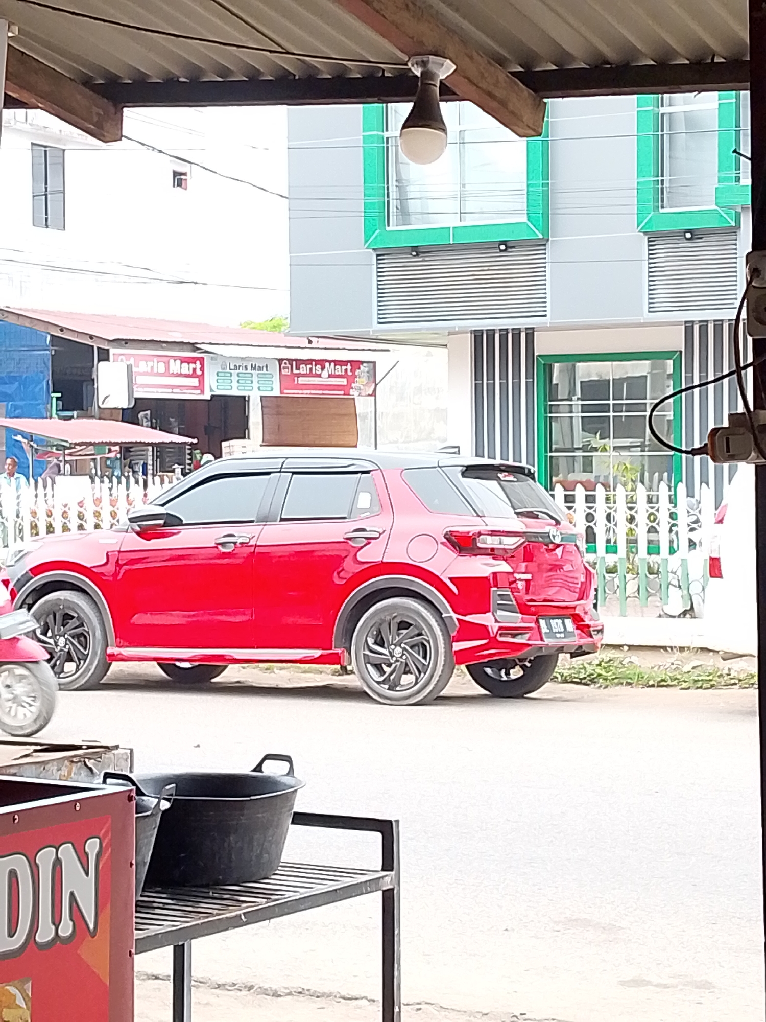 The image depicts a street scene in Lhokseumawe, Indonesia, under a covered structure. In the foreground, a bright red compact SUV is parked, its passenger side facing the viewer. The vehicle has tinted windows and black accents on the roof and wheel arches. To the left of the SUV, a portion of a red scooter is visible. In the background, a modern building with gray and green elements stands, featuring large windows and horizontal metal roll-up doors. A white picket fence runs along the front of the building. To the left of the building, a row of shops is visible, with signs indicating "Laris Mart." A man in a light-colored shirt and pants is standing near one of these shops. The ground is paved, and some greenery is present near the fence. A metal shelf with two large black plastic basins is positioned in the lower right of the frame, partially obscuring the road. A red sign with white lettering that appears to say "DIN" is visible on the left side of the image.