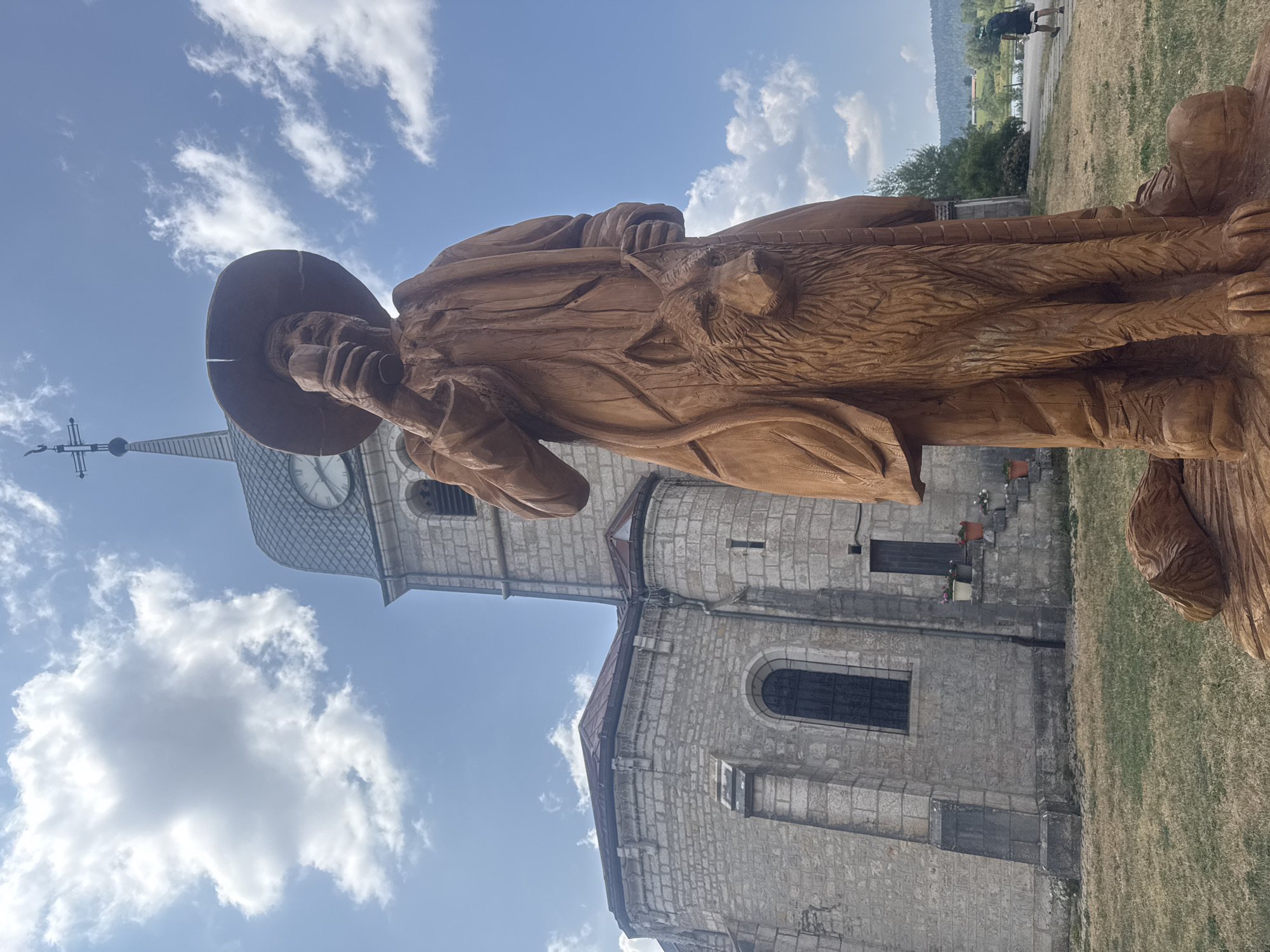 An outdoor, eye-level, full-shot photograph features a large, intricately carved wooden sculpture of a man wearing a wide-brimmed hat and a long coat, holding a glass in his right hand. A wolf is carved and integrated into the lower portion of the sculpture, appearing to be part of the man's cloak. The sculpture stands in a grassy area in front of a stone church with a steeple and a clock face. The sky above is blue with scattered white clouds. In the background, a paved path leads away from the church, with a person walking in the distance. The location is identified as Grande-Rivière Château, France.