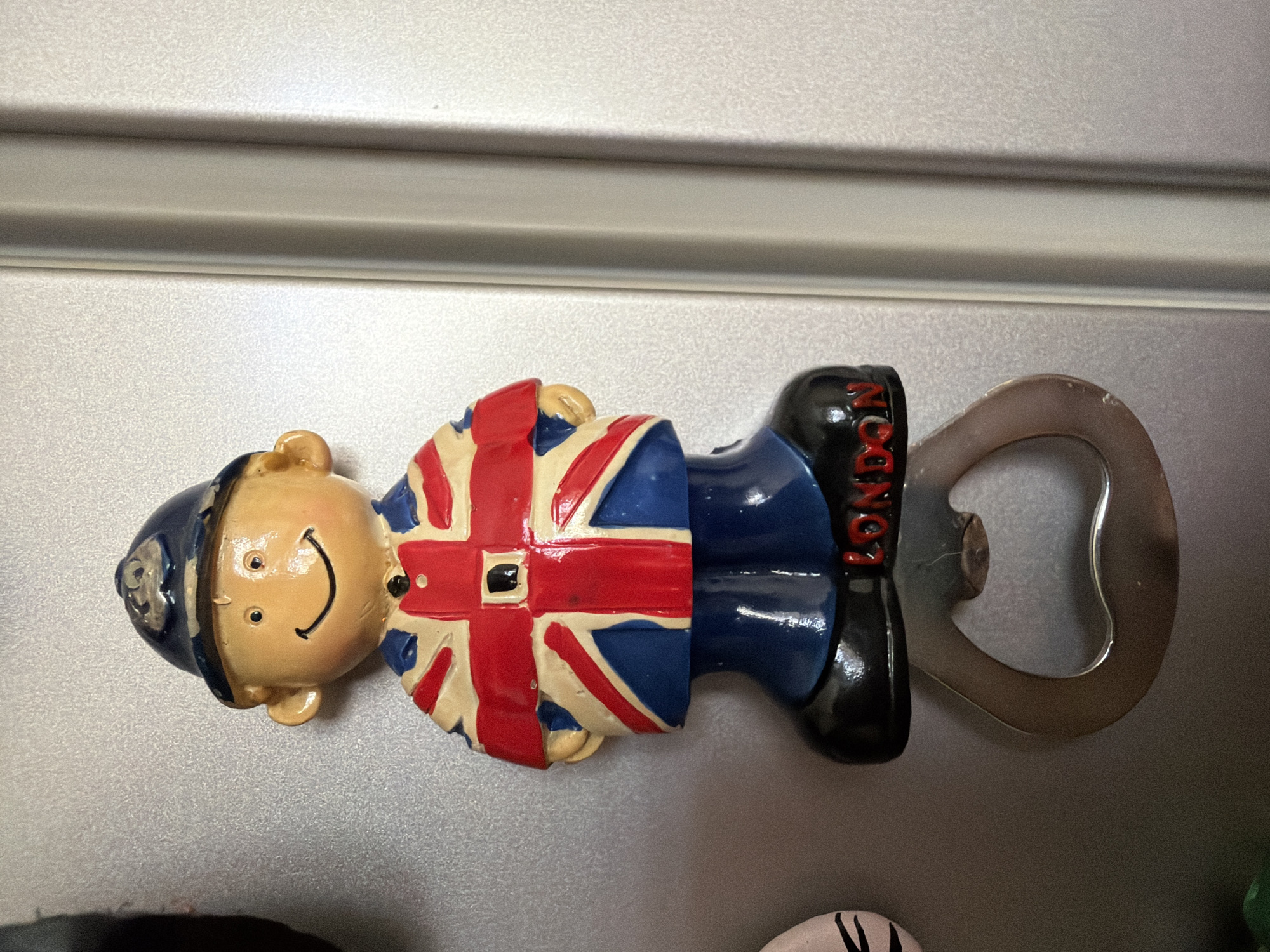 The image shows a novelty bottle opener shaped like a cartoon caricature of a British person, possibly a police officer, wearing a blue helmet and a uniform decorated with the Union Jack flag. The figure has a smiling face. Attached to the figure's leg is a metal bottle opener. The figurine is attached to a metallic surface, likely a refrigerator, as indicated by the horizontal ridges above the object. The background is a neutral, textured metallic surface. The scene appears to be indoors, with artificial lighting. There is some text visible on the figure's boot, which appears to read "LONDON". The image is taken from a slightly elevated angle, with the figure oriented horizontally.