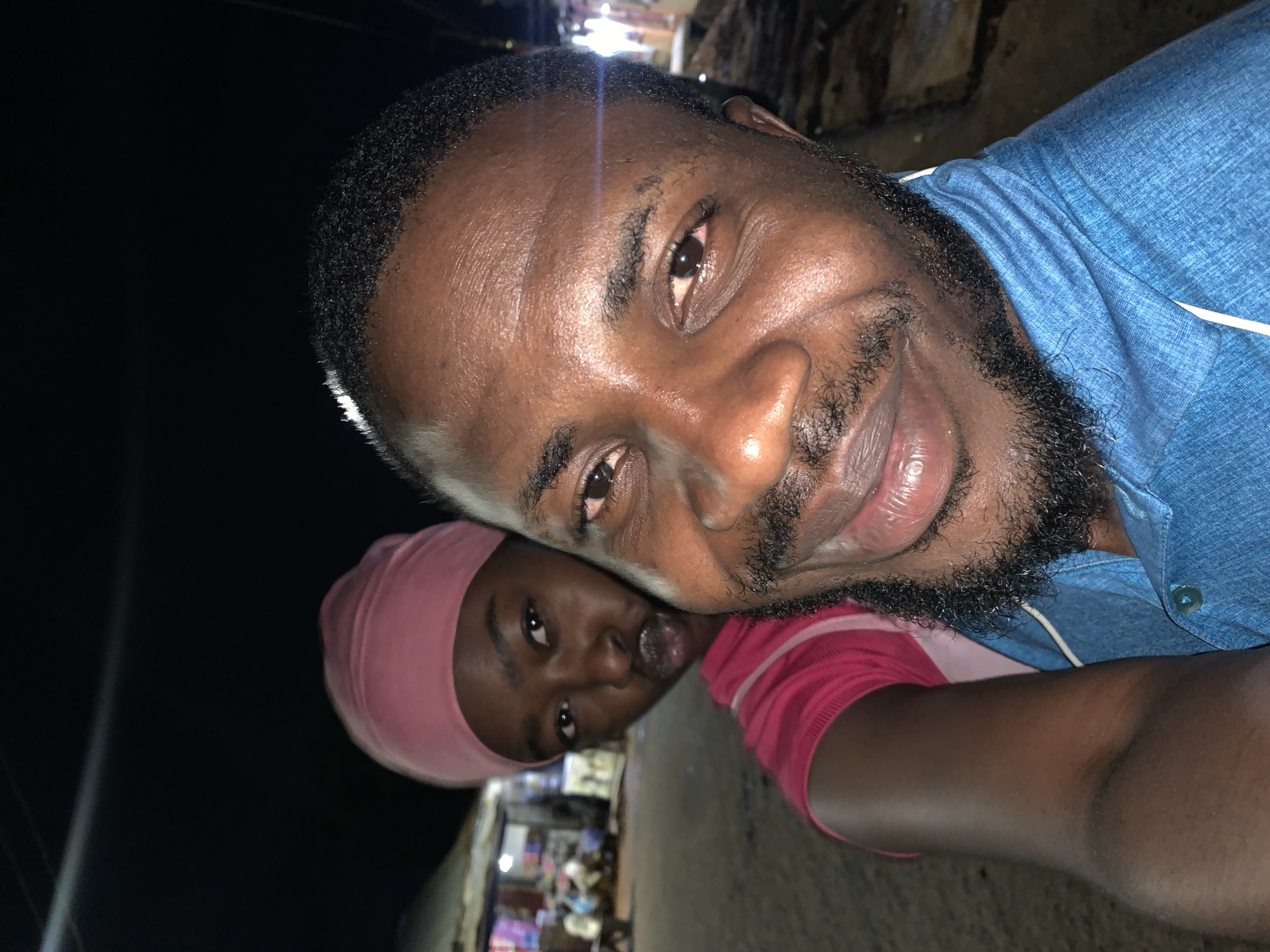 The image shows a selfie taken by a dark-skinned man with a close-cropped haircut, a beard, and a blue shirt. He is smiling at the camera. Behind and to the left of the man is a dark-skinned woman wearing a pink head covering and a pink and white striped shirt. They appear to be standing in a lit area, possibly a street or alley. In the background, there are structures and people, suggesting they are in a public area. The location appears to be in Kabong, Nigeria.
