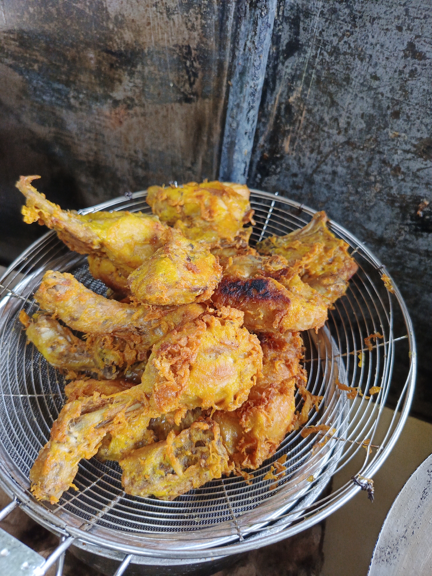 A pile of freshly fried chicken pieces rests in a metal mesh basket, likely a frying basket, set against a backdrop of textured, aged surfaces. The chicken pieces vary in shape and size, some appearing to be wings and drumsticks, with a deep golden-yellow color indicating they have been deep-fried. The texture suggests a crispy coating. The basket has a concentric circular pattern of metal mesh. The background has a mottled appearance, with dark, uneven areas possibly indicating aged concrete or plaster. The setting suggests an outdoor or semi-outdoor cooking area. The location is Lhokseumawe, Indonesia.