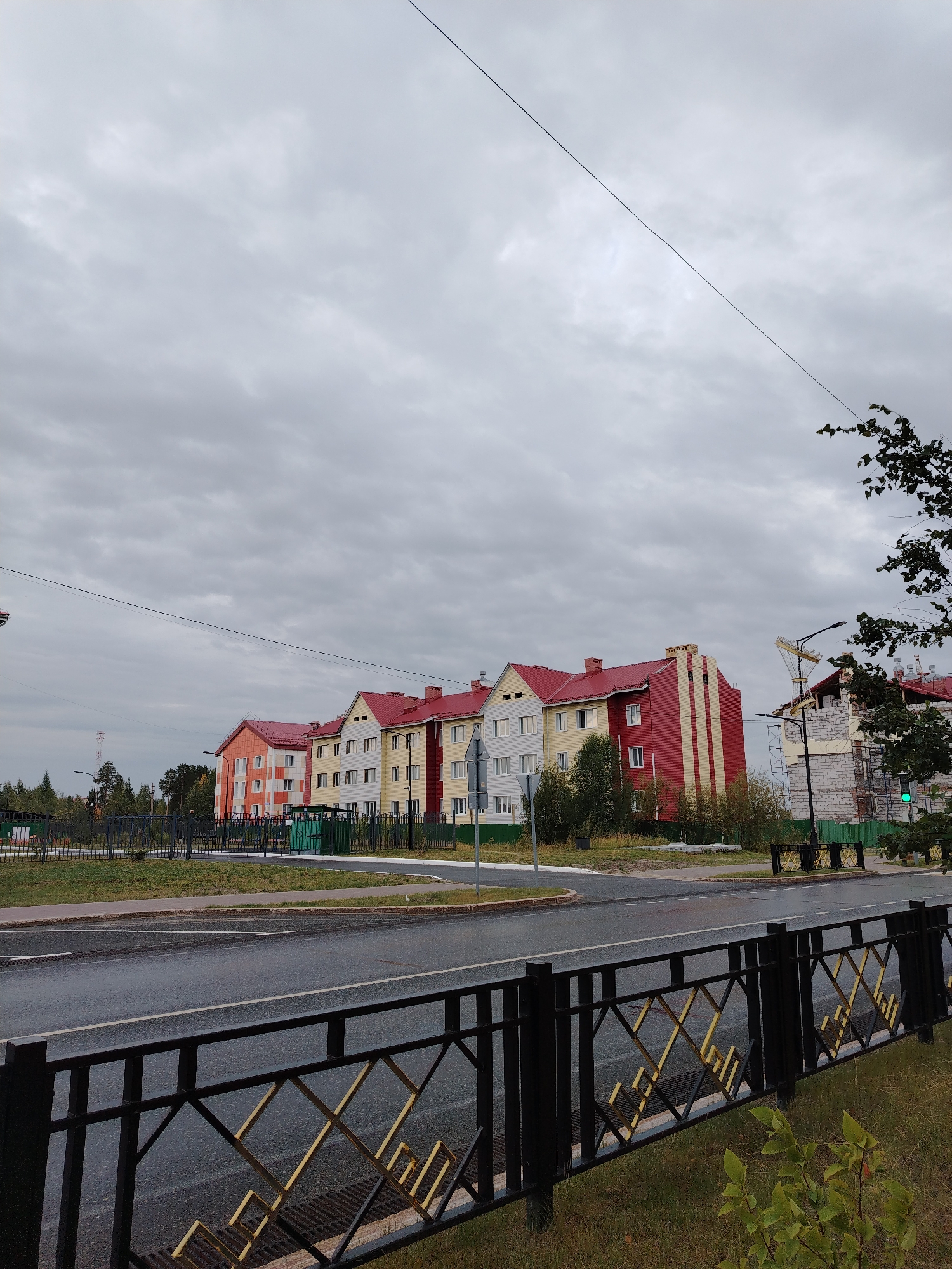 The image shows a row of apartment buildings with red roofs and multi-colored facades, located in the city of Muravlenko, Russia. The buildings are of varying heights and architectural styles. Some are finished with yellow, orange, and white panels, while others are still under construction with visible brickwork and scaffolding. In the foreground, a wet asphalt road is visible, separated by a black metal fence with decorative yellow elements. A green traffic light is visible on the right side of the image, indicating an intersection. The sky is overcast with grey clouds. A tall lattice communication tower is visible in the distance. The overall scene depicts a residential area under development on a cloudy day.