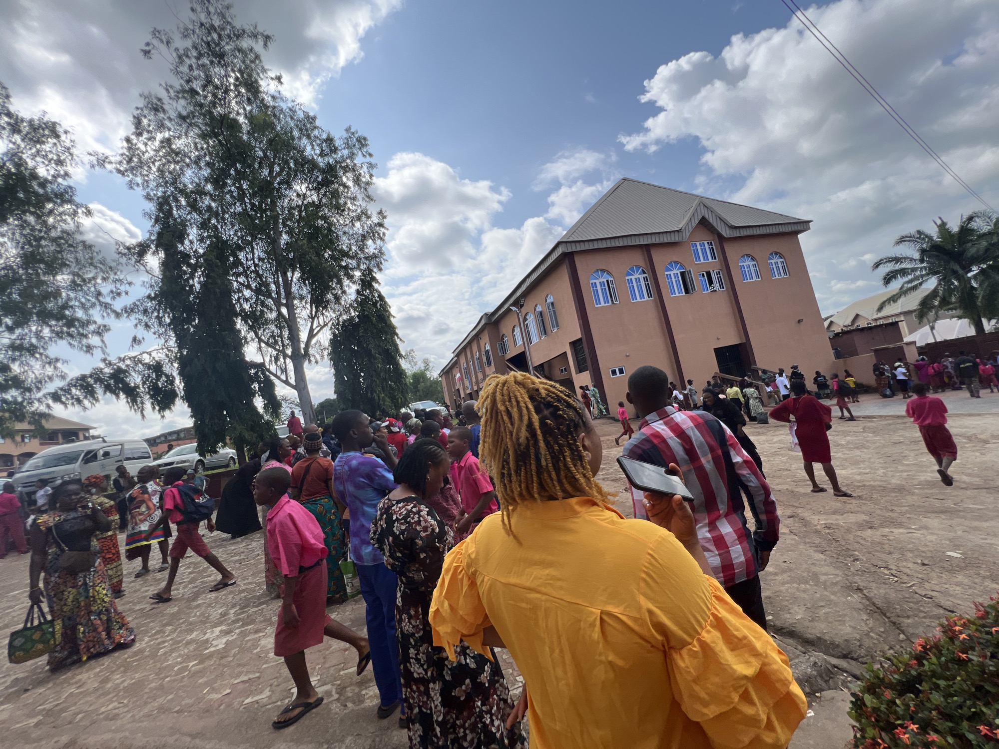 The image captures a lively outdoor gathering in Nnewi, Nigeria, likely in front of a modern, multi-story building with a beige facade and a grey, gabled roof. The building features numerous arched windows with blue panes and white trim, suggesting it could be a school, church, or commercial establishment. A diverse crowd of people, dressed in colorful attire, fills the foreground and middle ground. Many individuals appear to be engaged in conversation or observing the scene, with some children in school uniforms, including a boy in a bright pink shirt and shorts, and a girl in a pink shirt and skirt. One woman in the foreground wears a vibrant orange shirt with puffy sleeves and has her hair styled in blonde dreadlocks, holding a smartphone. Other individuals are seen in traditional Nigerian fabrics. A red vehicle, possibly a van, is parked on the left side of the image, and in the background, other buildings and palm trees are visible. The sky is partly cloudy, with fluffy white clouds against a bright blue sky. The ground is paved with a reddish-brown surface. The overall atmosphere suggests a community event or a school function taking place on a sunny day.