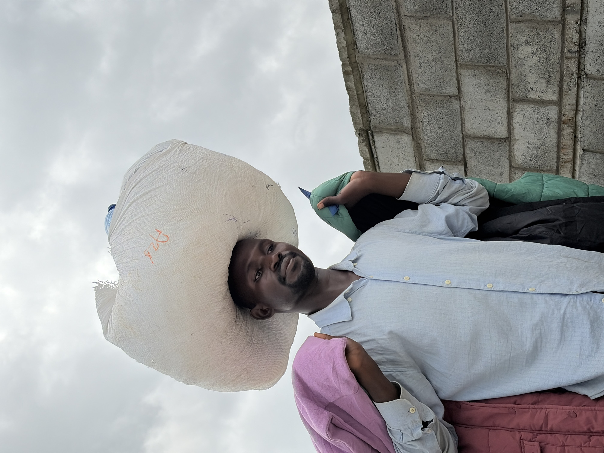 A dark-skinned man with a short beard stands outdoors, carrying a large white sack balanced on his head. He is wearing a light blue button-down shirt. Draped over his left shoulder is a folded pink garment, and over his right shoulder is a green padded jacket. To his left, a dark red quilted jacket is visible. Behind him, a wall constructed of grey concrete blocks extends horizontally. The sky above is overcast and cloudy. The scene appears to be in Igboefon, Nigeria, based on the context provided.