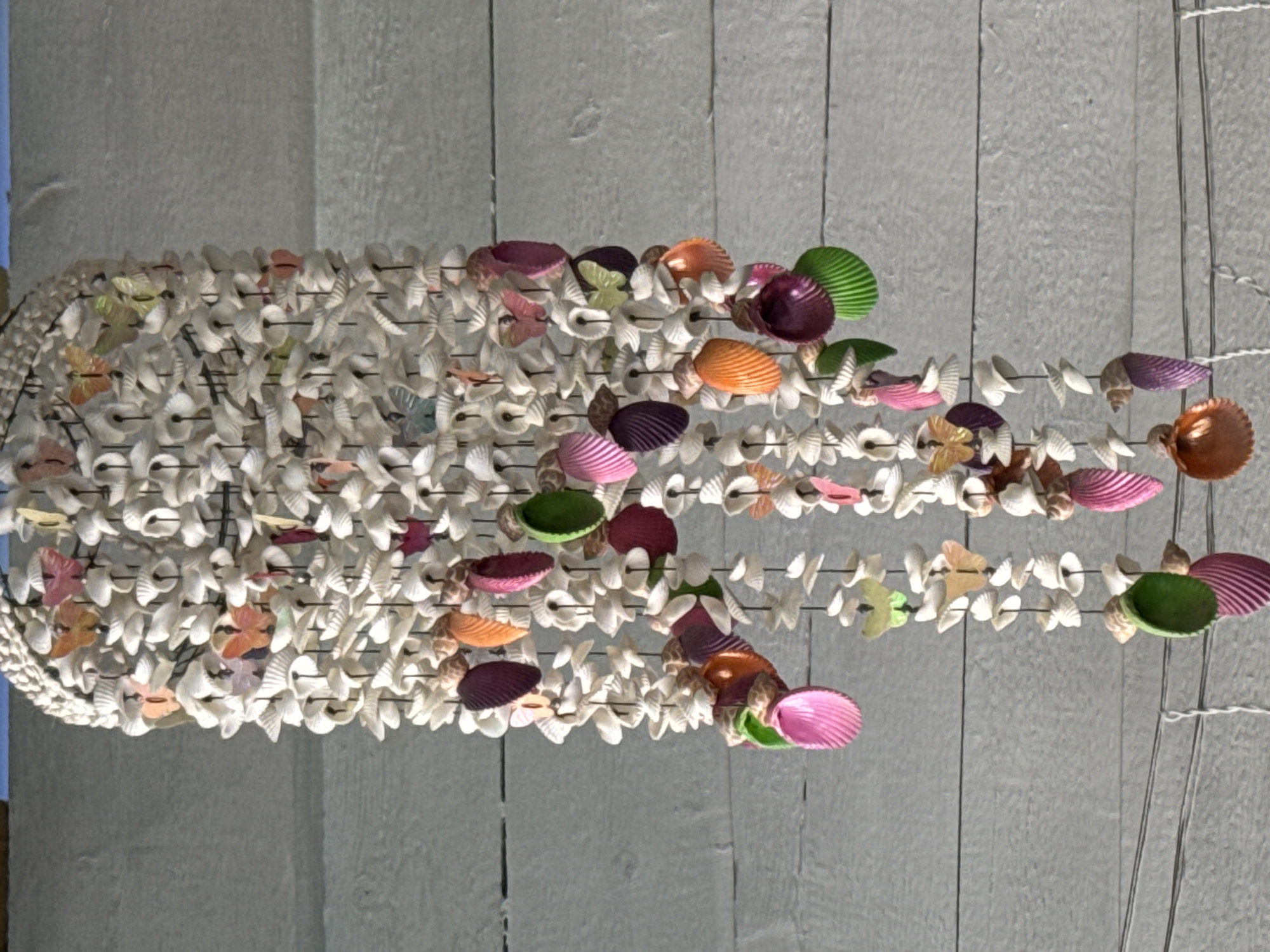 A decorative mobile made of small white seashells and colorful, butterfly-shaped ornaments hangs from wires against a weathered, light gray wooden wall. The mobile is a cascading structure with multiple strands of shells and beads, interspersed with bright accents of pink, orange, green, purple, and yellow. The butterfly ornaments are primarily located towards the top of the mobile and are visible in various colors and sizes. The wooden wall behind the mobile features horizontal planks and shows signs of age and wear. The scene is outdoors, suggested by the natural lighting and the background wall, which could be part of a building or fence. There are no people or animals visible in the image.