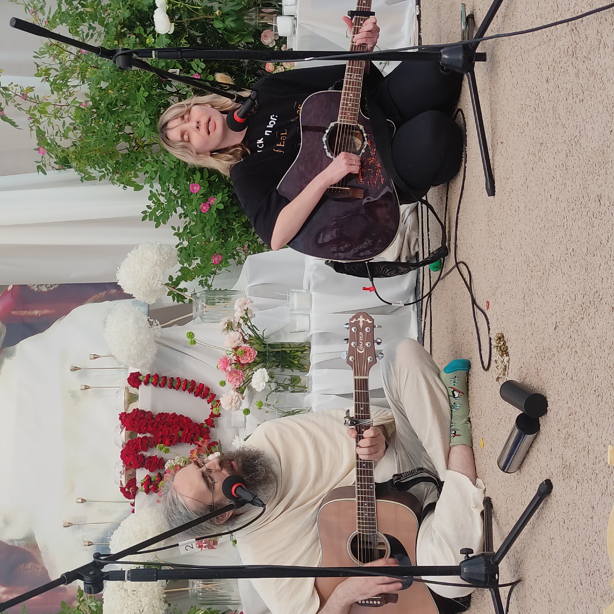 The image depicts an outdoor gathering, possibly a festival or performance, in the city of Zhirnosovo, Russia. Two individuals are seated on a carpeted floor, each playing an acoustic guitar. The person on the left, a man with a beard and glasses, wears a light-colored robe and plays a tan acoustic guitar. He is positioned in front of a microphone stand. To his right, a woman with blonde hair, wearing a black t-shirt, plays a darker acoustic guitar and is also positioned near a microphone stand.
The background suggests a decorated setting. A white cloth covers a raised surface behind the musicians, adorned with garlands of red flowers and smaller bouquets of pink and white flowers in vases. Two tall white floral arrangements are also visible. Behind the floral arrangements, white curtains create a backdrop, and a large image, partially obscured, hangs on the left. Green foliage with pink flowers is present on the right side of the frame, suggesting a garden or natural setting.
On the carpet in front of the musicians, several objects are scattered. These include two cylindrical metal objects, a set of small bells on a string, and a guitar pick. The overall atmosphere appears to be one of a peaceful musical performance.