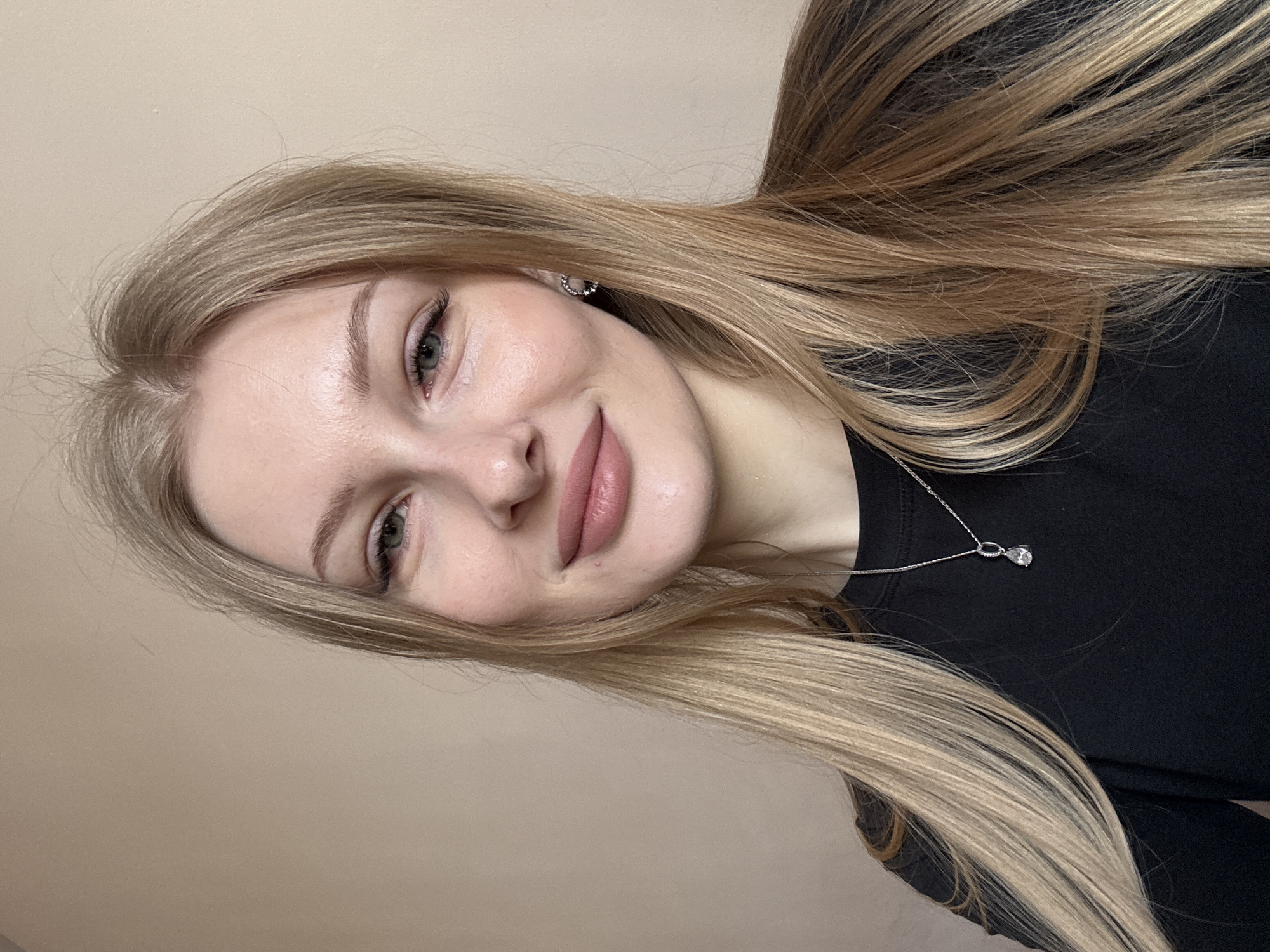 An image taken indoors in Perm, Russia, depicts a young adult female from the chest up, framed against a plain, light beige wall. The subject has long, straight blonde hair with lighter highlights, parted slightly to her left, flowing over her shoulders. Her face is oval, with fair skin, light-colored eyes (appearing green/hazel), and visible eyelashes and defined eyebrows. She has full lips with a light pink/nude shade and a small mole above the right side of her upper lip. She is exhibiting a soft, pleasant smile. She wears a black, long-sleeved top. Adornments include a silver-colored chain necklace with a clear, teardrop-shaped pendant positioned centrally on her chest, and a silver-colored stud earring in her right ear, appearing as a small cluster or curved design. The lighting is even and soft.