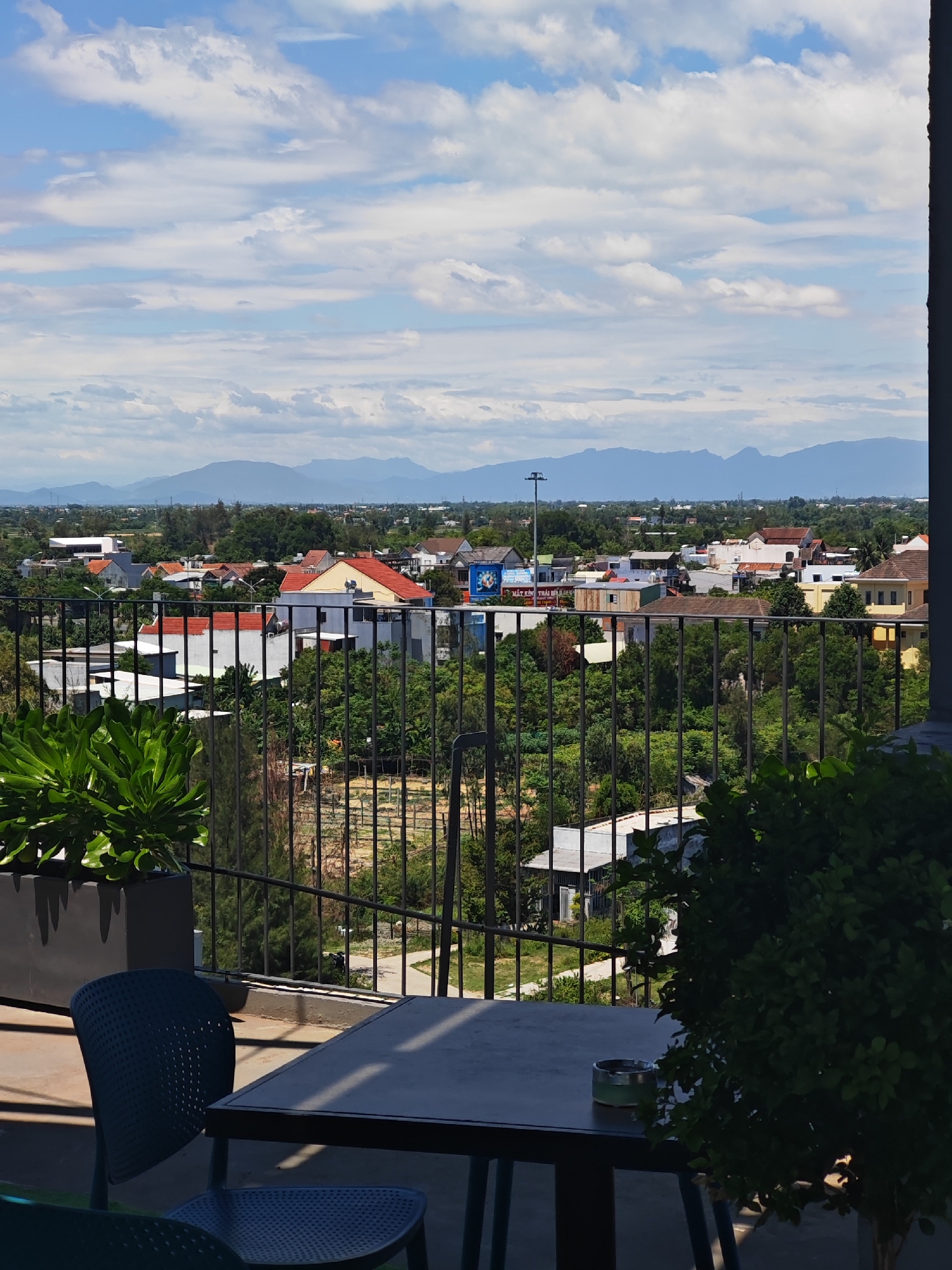 This is a landscape photograph taken from a high vantage point, likely a balcony or rooftop, overlooking a city in Đà Nẵng, Vietnam. The foreground features a table with an ashtray, two chairs, and a large potted plant on the left, all casting shadows on a concrete floor. A black metal fence separates the seating area from the view beyond.
The midground presents a dense canopy of green trees, interspersed with various residential buildings, some with red tiled roofs and others with white or yellow facades. A tall streetlamp is visible in the distance, along with power lines and utility poles.
The background is dominated by a range of distant mountains under a bright blue sky with scattered white clouds. The overall scene suggests a partly cloudy day with good visibility. The presence of trees and buildings indicates an urban or suburban environment. The architectural styles of the buildings, particularly the red tiled roofs, are characteristic of Southeast Asian urban areas. The ashtray on the table suggests a public or semi-public outdoor space, possibly a cafe or restaurant.