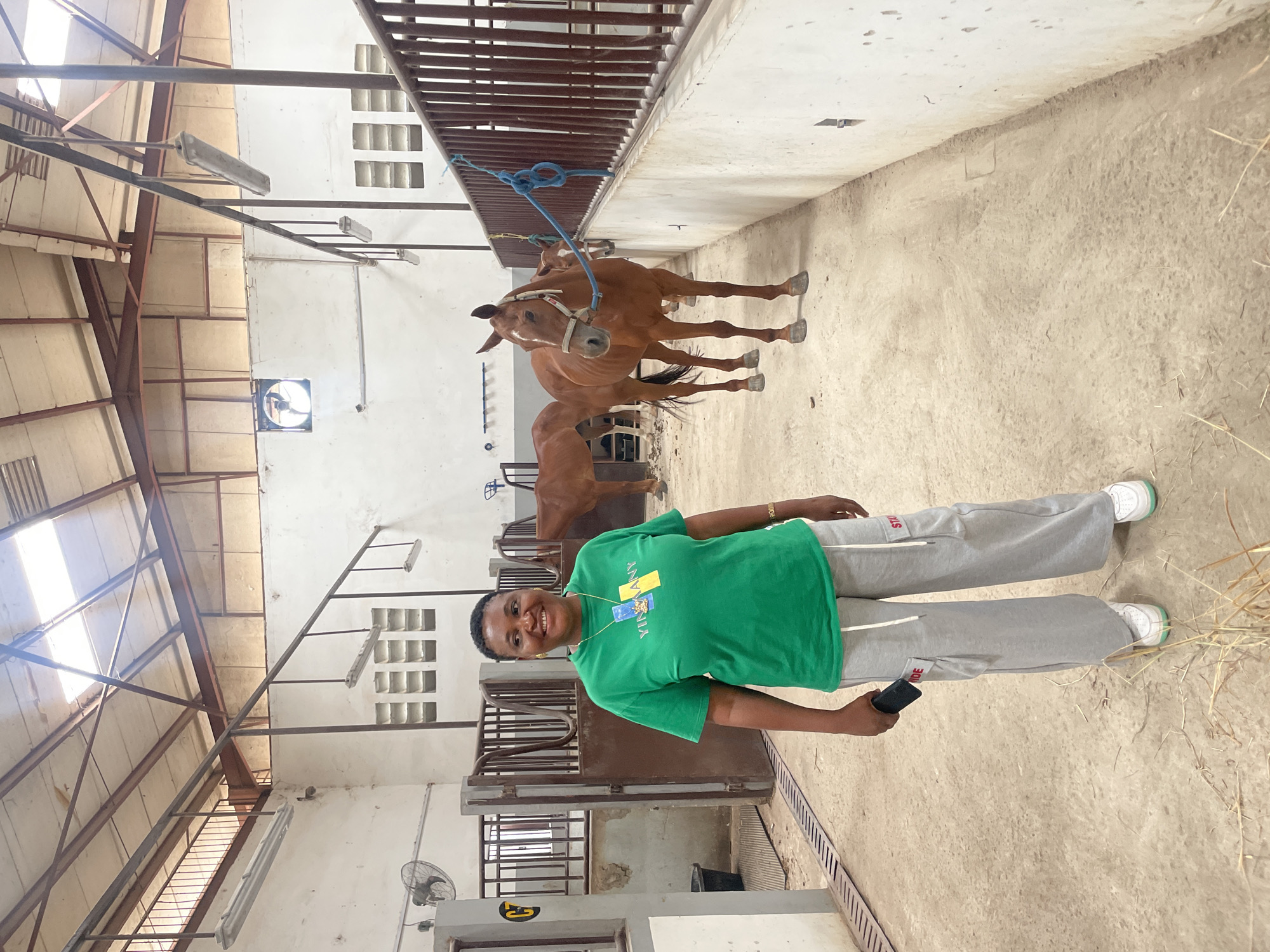 The image shows an indoor scene, likely a stable or barn, in Hayin Banki, Nigeria. A woman stands in the foreground, wearing a green t-shirt with yellow patches and grey sweatpants with white sneakers. She holds a dark object, potentially a mobile phone, in her right hand.
Behind her, two chestnut-colored horses are visible in separate stalls. The stalls are constructed with dark metal bars. A blue rope is tethered to the front of the stall containing the horse closest to the camera.
The environment consists of a light-colored concrete floor, with straw scattered on the ground near the woman. The walls and ceiling are also light-colored. There's a metal ceiling structure. A ventilation fan is present on the upper part of the wall. Light fixtures are attached to the ceiling.