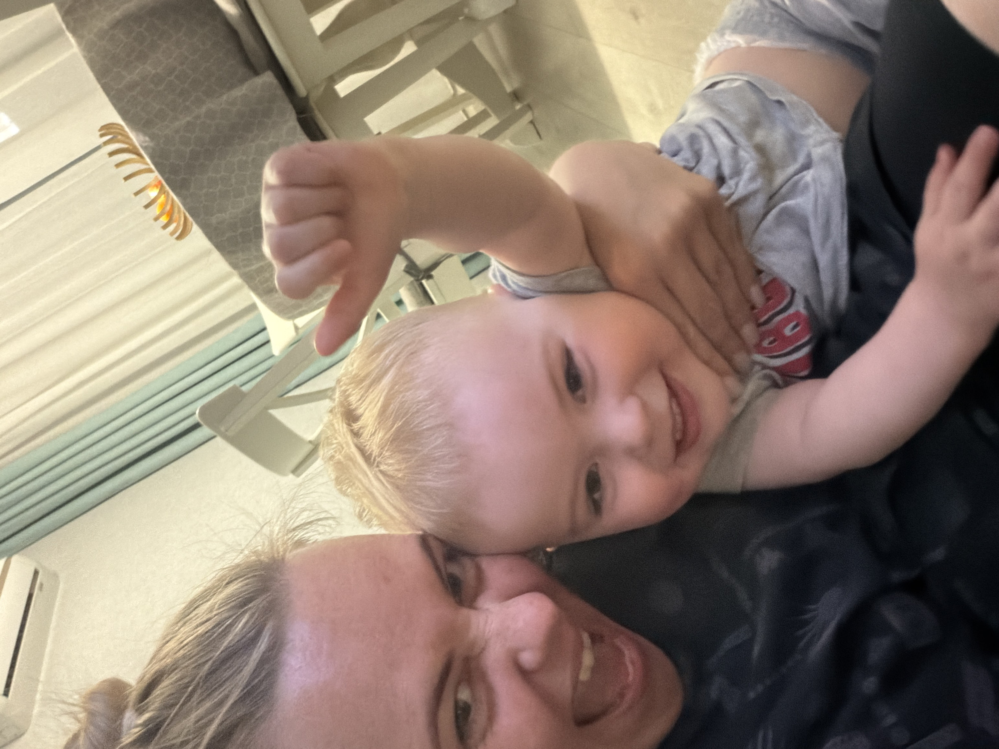 A selfie image shows a blonde woman with light skin and a baby with blonde hair. The woman is smiling broadly with her mouth open, revealing her teeth. The baby is also smiling and appears to be lying on top of the woman. The baby is giving a thumbs-down with one hand. The background includes a slatted blind, a white wooden chair or crib, and an air conditioning unit. There is no information in the image to suggest that it was taken in the city of Votkinsk, Russia.