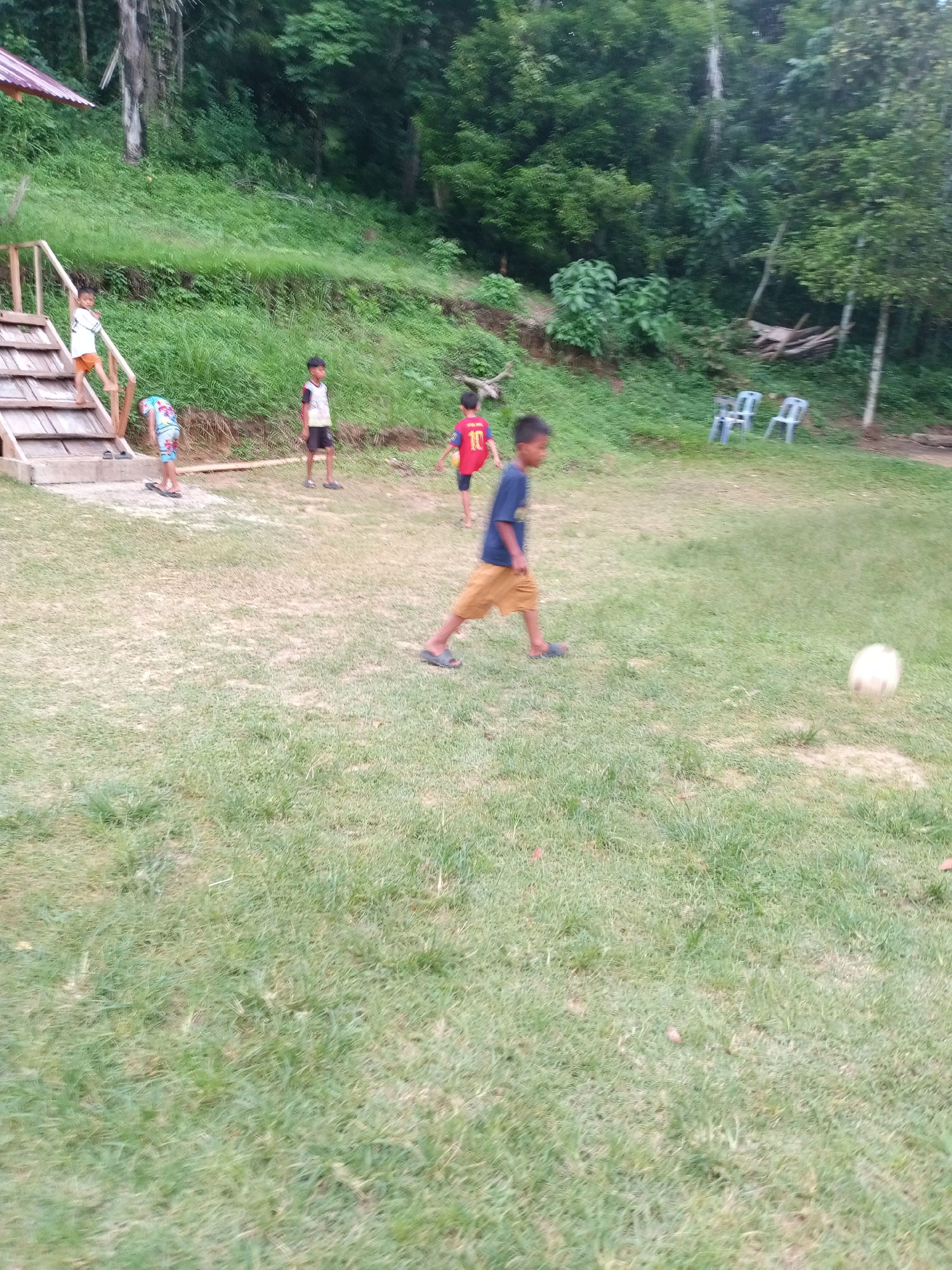 The image depicts an outdoor scene with several children playing in a grassy area. In the foreground, a young boy in a blue shirt and brown shorts is running towards a soccer ball, which is visible in the mid-right of the frame. To the left, another boy in a red shirt with the number '10' on it is standing, holding a soccer ball. Further to the left, two more boys are visible near a wooden staircase. One boy is standing at the top of the stairs wearing an orange shirt, and another is bending down near the bottom in a brightly colored patterned outfit. In the background, a dense forest with lush green trees covers a sloping hillside. There are a few plastic chairs scattered in the middle ground, suggesting a communal or recreational area. The setting appears to be a rural or semi-rural location, possibly a park or an open field adjacent to a wooded area, with a small structure visible in the upper left corner, likely a shelter or part of a building. The sky is not clearly visible due to the foliage. The overall impression is one of children engaged in outdoor play in a natural environment.