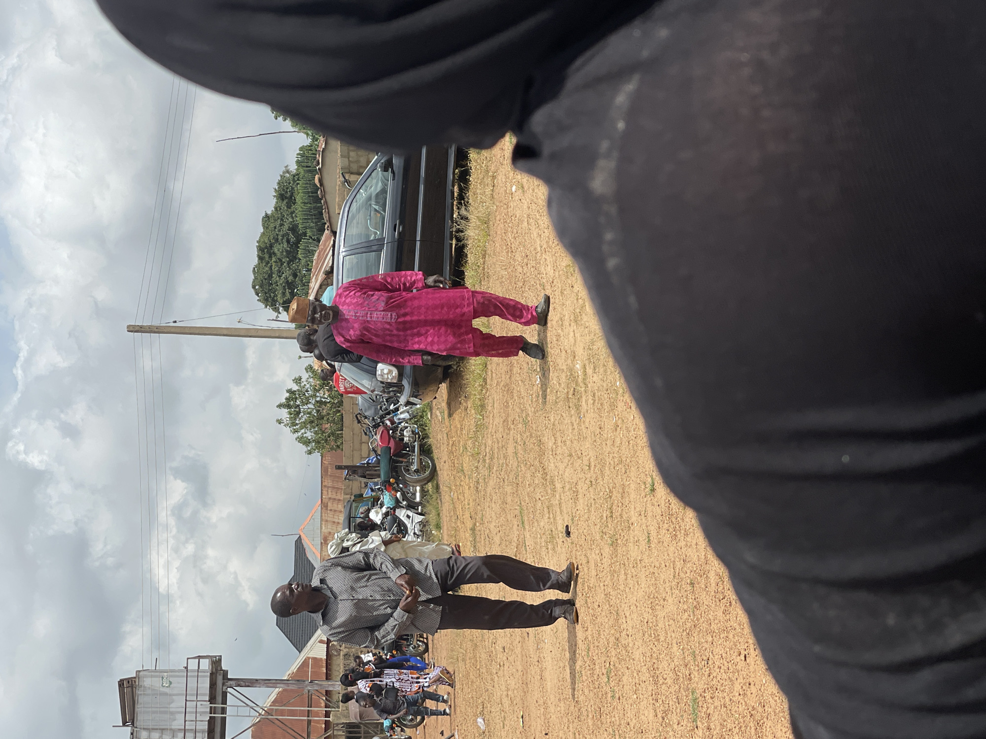 The image captures an outdoor scene in Kassa, Nigeria, under a cloudy sky. In the foreground, a person wearing a black hijab is partially visible, facing away from the camera. Two men are the main subjects in the midground. The man on the left is wearing a grey patterned shirt and dark pants, looking towards the left of the frame. The man on the right is dressed in a vibrant pink traditional outfit and a matching cap, with his gaze directed forward. Behind them, a black sedan is parked, and several motorcycles are visible to the left of the car. In the background, buildings with corrugated iron roofs and a water tower are present, along with trees and a utility pole. The ground is covered in loose, sandy soil. The overall atmosphere appears to be that of a public gathering or event.