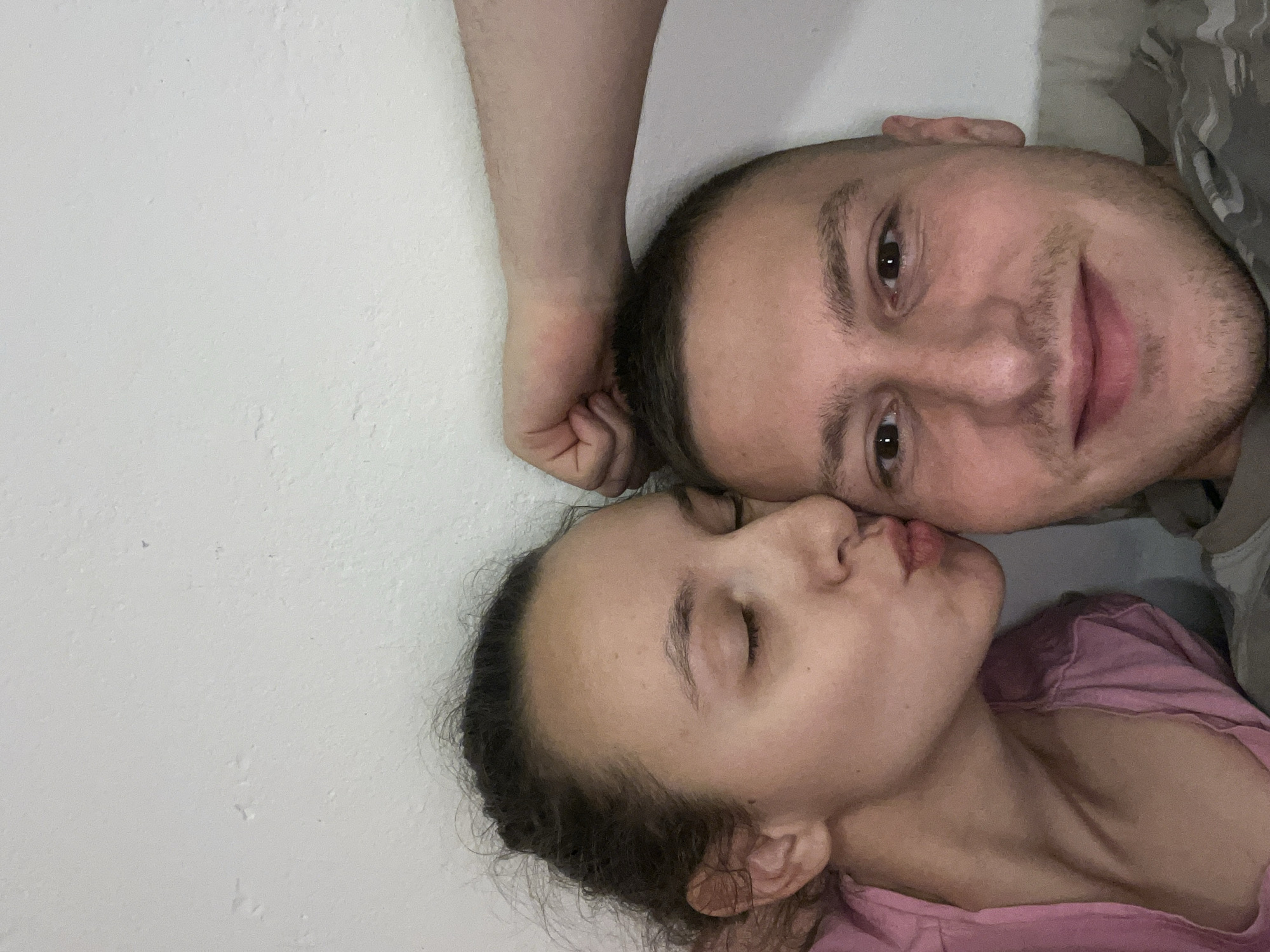 A close-up selfie shows a light-skinned male and female lying down. The man is to the right, smiling with dark eyes, short dark hair, and stubble. He's wearing a gray camo shirt. The woman, wearing a pink shirt, is on the left with her eyes closed and lips puckered, as if giving a kiss. A white wall occupies the top and left of the frame. The photo's location in Saarbrücken, Germany, is unknown.