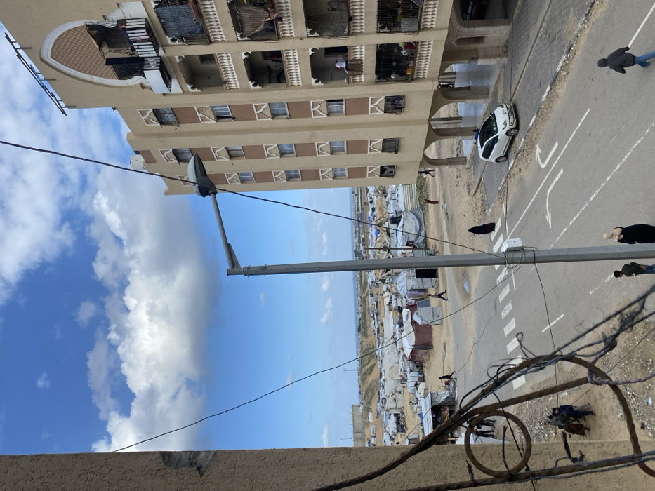 This elevated view in Khan Yunis, Palestinian Territory, captures a street scene bordered by a multi-story residential building and a sprawling temporary encampment, under a partly cloudy blue sky.
In the foreground, numerous dark, tangled wires crisscross the frame from the upper left. A gray street light pole stands centrally, featuring a single luminaire. Beneath, a paved road includes a white crosswalk and a white left-turn arrow marking. A white compact car, possibly a Volkswagen Up!, is parked or moving slowly on the right side of the road, facing left.
Multiple individuals are visible on the street: a person in a black abaya walks across the crosswalk, two individuals stand near the street pole (one with a light head covering), another person in dark clothing runs towards the lower right, and a group of approximately five people are gathered on the far left side of the road near a makeshift stall.
To the left of the road, a vast temporary encampment stretches towards the distant horizon, characterized by numerous tents and improvised shelters constructed from tarpaulins, fabrics, and other materials, appearing in various shades of white, gray, and brown. Several individuals are observed walking within this encampment, which includes what appears to be a small market stall under a blue tarp near the road. Beyond the encampment, a large, light-colored elevated structure, possibly a water tank, is visible, and the faint line of the sea is discernible on the horizon.
To the right of the road stands a multi-story (at least seven visible floors) residential building with a beige facade, vertical brown decorative panels, and arched windows and balconies. Clothes and tarps hang from multiple balconies, and at least three individuals are visible on different balconies, including one man in a white shirt on a higher floor. The ground level of this building is supported by open arches, creating an open space beneath.