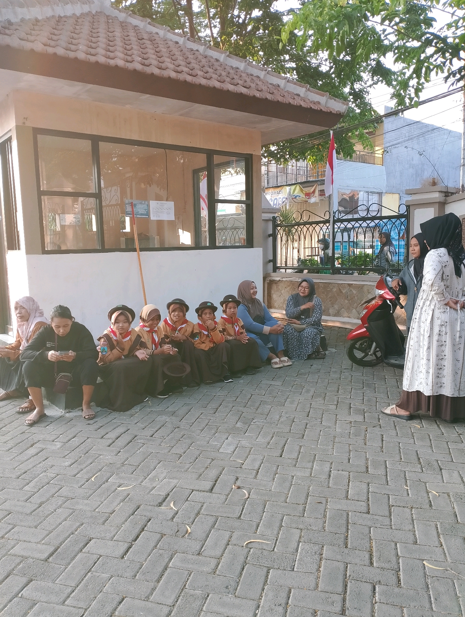In Probolinggo, Indonesia, a group of young scouts in brown uniforms and hats are sitting on a paved courtyard in front of a low-rise building. The building has a tiled roof and large windows. Several adult women are also present, some sitting with the children, while others stand near a red scooter. One woman is looking at her phone. The scene appears to be an outdoor gathering or waiting area, possibly associated with an educational institution or community center, given the presence of the scouts and the building's appearance.